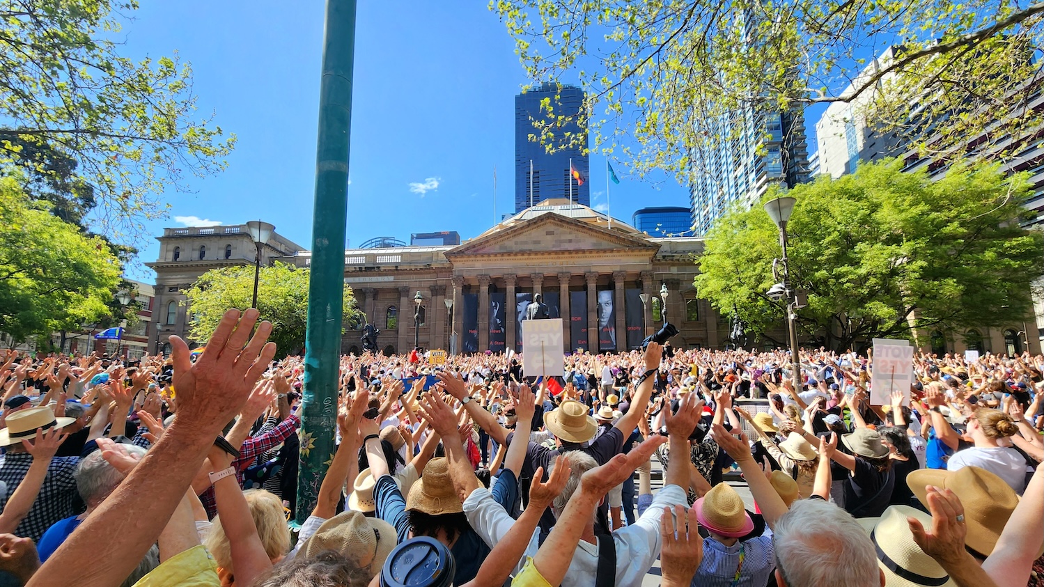 Large crowd gathered at a Yes23 campaign rally in Australia, with hundreds of people raising their hands in front of a historic civic building, advocating support for the Indigenous Voice to Parliament referendum under a clear blue sky.
