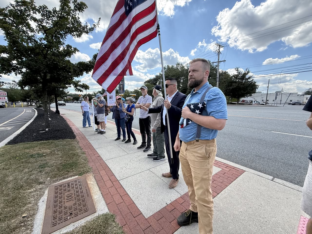 State assembly candidate Citron stands with community members holding an American flag during a roadside demonstration, engaging residents in civic participation.