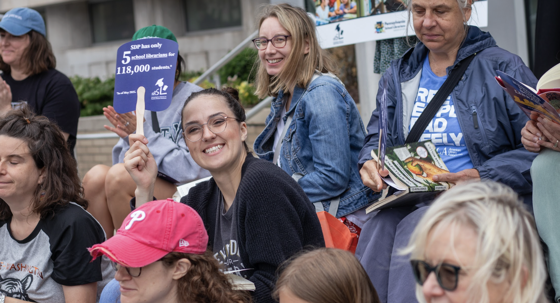 A crowd of rally attendees, mostly women, smile and hold signs at an outdoor library advocacy event. A woman in the center holds a purple paddle sign reading "SDP has only 5 school librarians for 118,000 students.* *As of May 2025." Visible branding includes the American Library Association and Pennsylvania Association for School Librarians (PASL). One attendee wears a blue shirt reading "FREE PEOPLE READ FREELY."