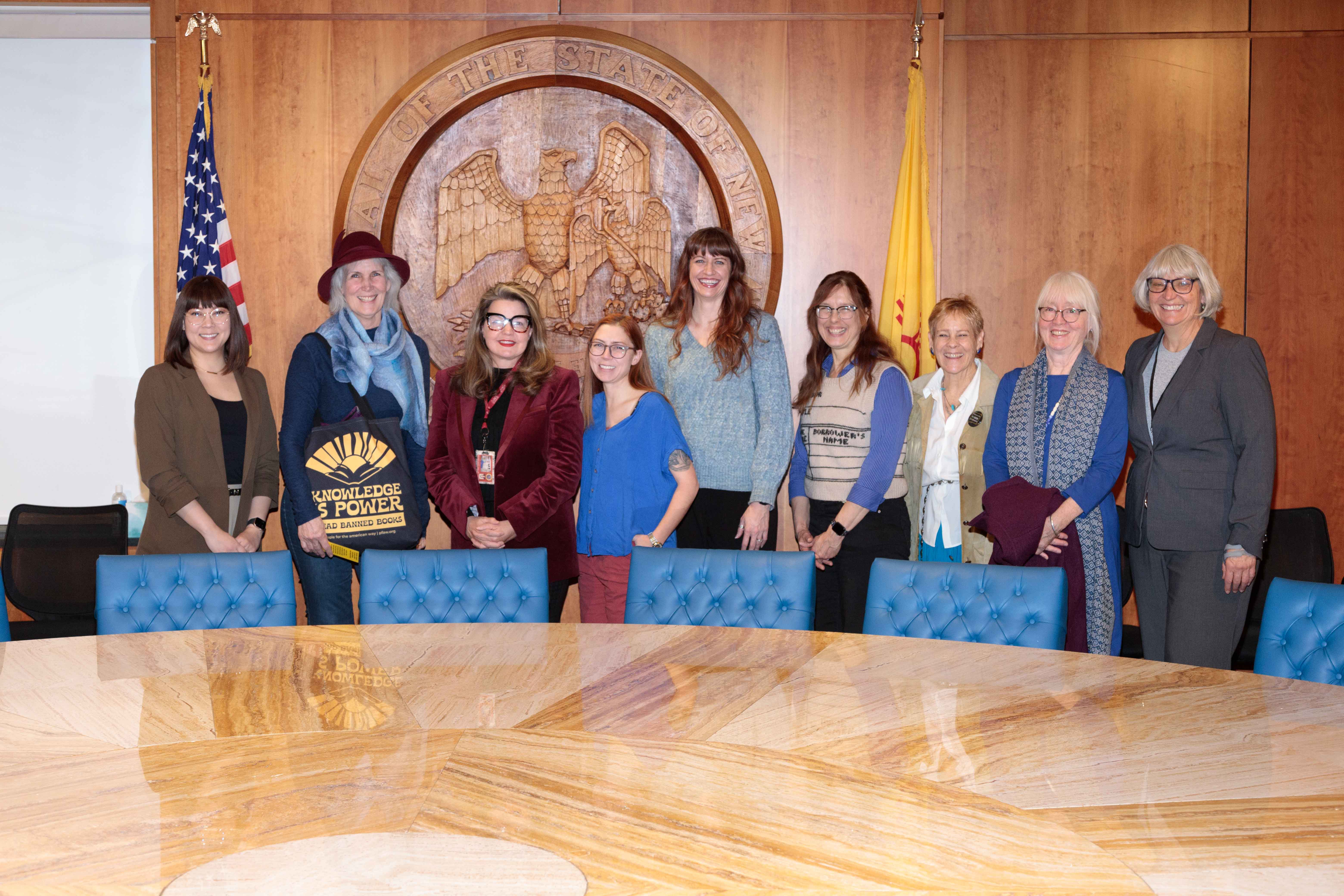Nine women stand in a formal conference room at the New Mexico State Capitol, posing in front of the carved state seal. A U.S. flag and New Mexico state flag flank the seal. One woman holds a tote bag reading "Knowledge is Power — Read Banned Books." Another wears a sweater with a library card design. A large marble conference table with blue leather chairs is in the foreground.