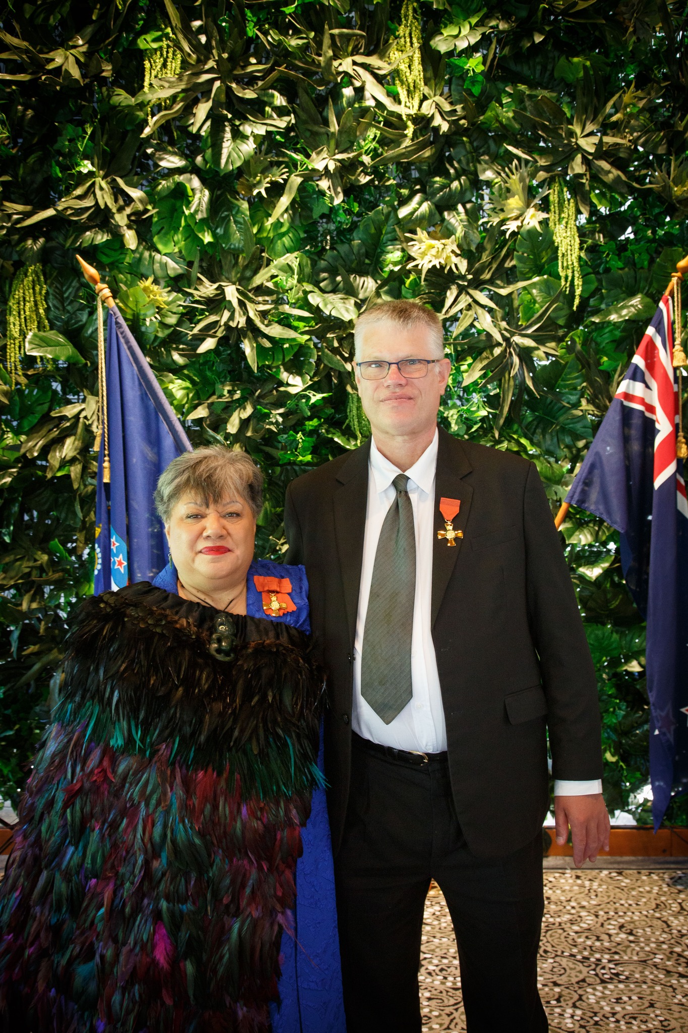 Chrissie Cowan wearing korowai and David Corner wearing black suit with ONZM medals, flags in backgound.jpg