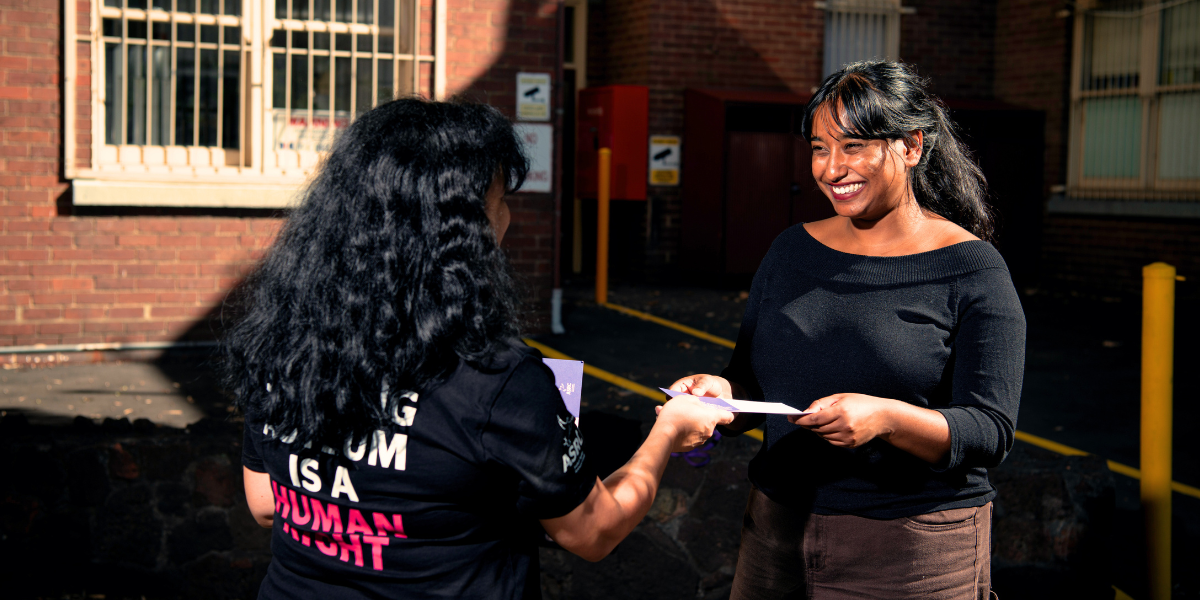 Two people, one with their back to the camera handing the other person a flyer. The person facing the camera is smiling.