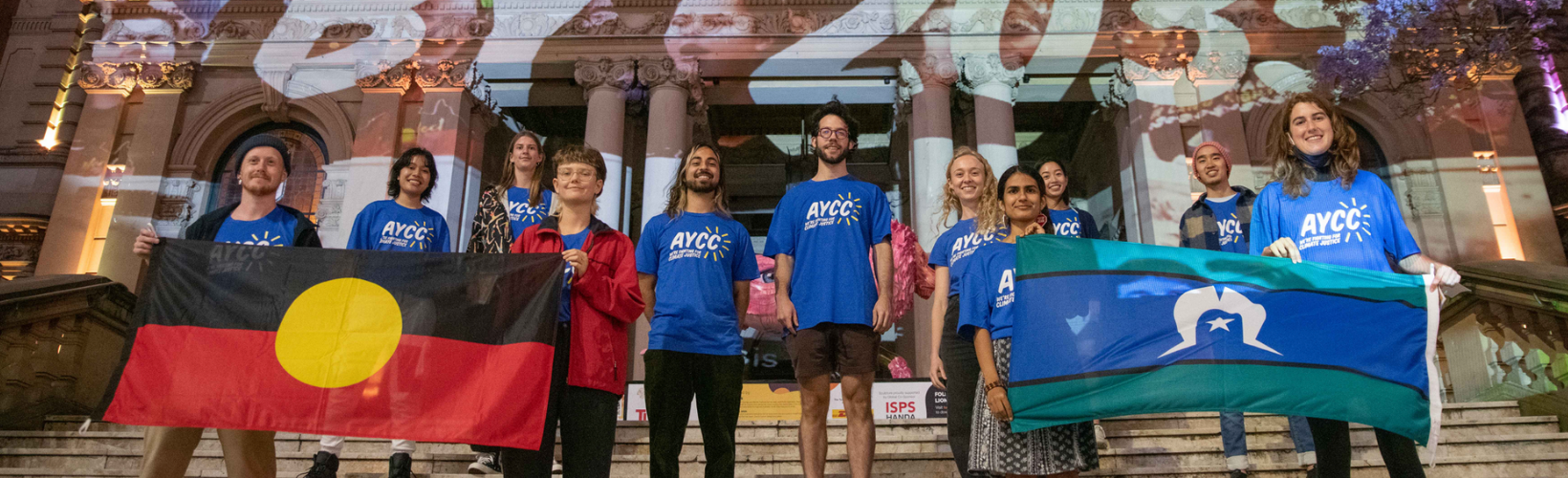 young people in blue AYCC shirts outside a projection onto Sydney town hall, holding up Aboriginal and Torres Strait Islander flags