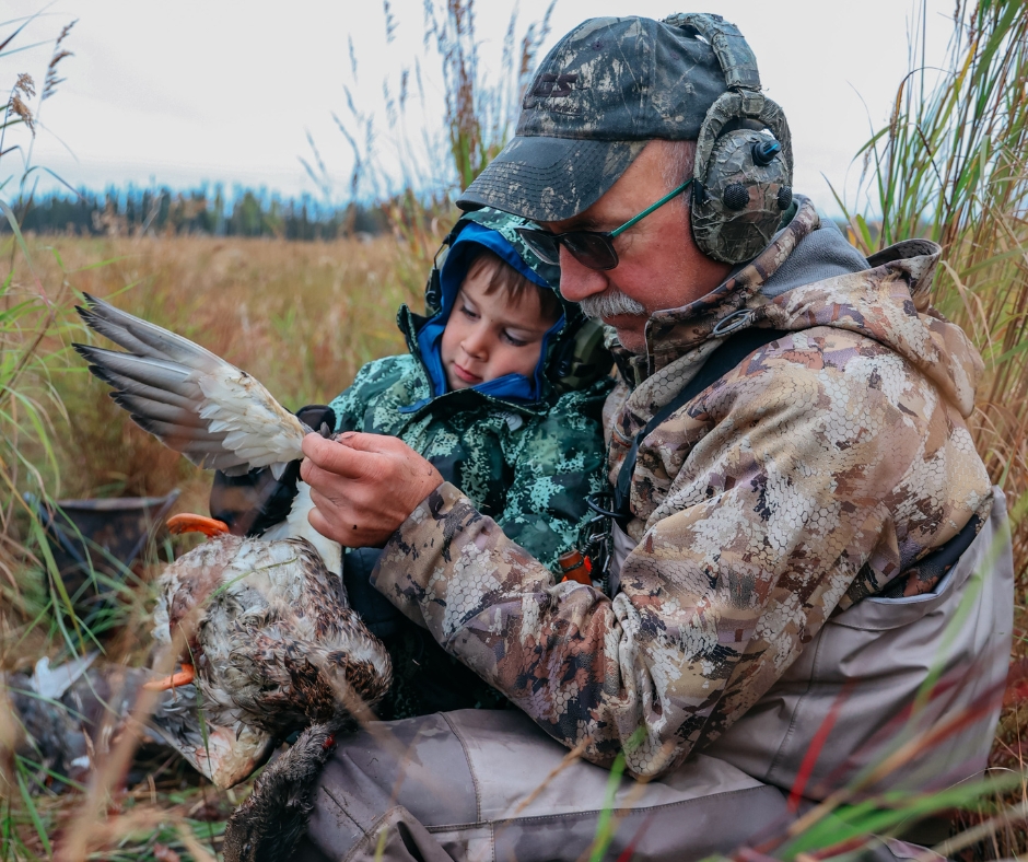 Grandfather and grandson sit in field, admiring duck they just hunted.
