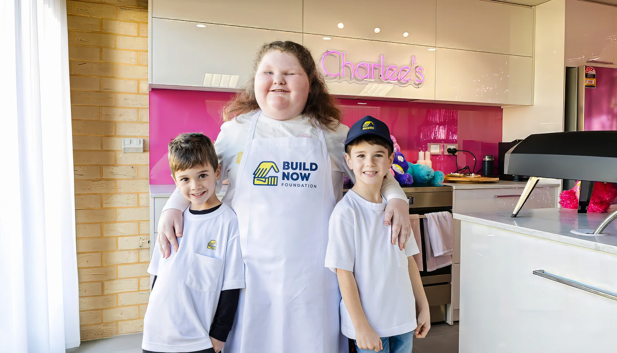 Charlee smiling in her newly built kitchen wearing a Build Now Foundation apron, a young boy either side of her, beneath a pink neon sign reading Charlee's