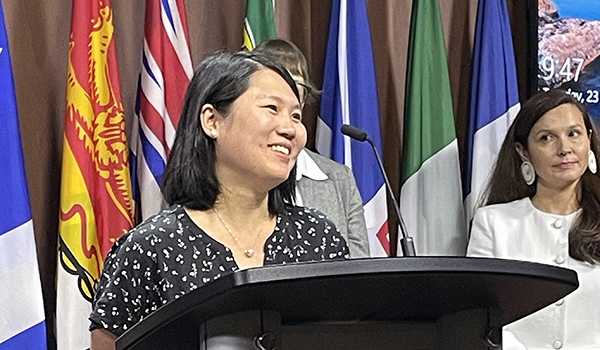 Dr. Helen Hsu smiling behind a podium with flags behind her