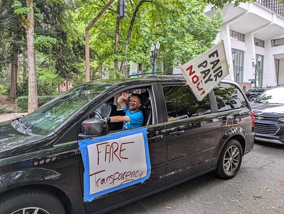 Rideshare driver in van circling the Portland Building in driver caravan with drivers's rights signs taped to the van.
