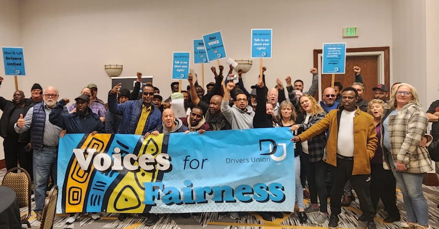 A large group of people holding a Drivers Union banner that reads "Voices for Fairness" hold signs and raise fists in the air.