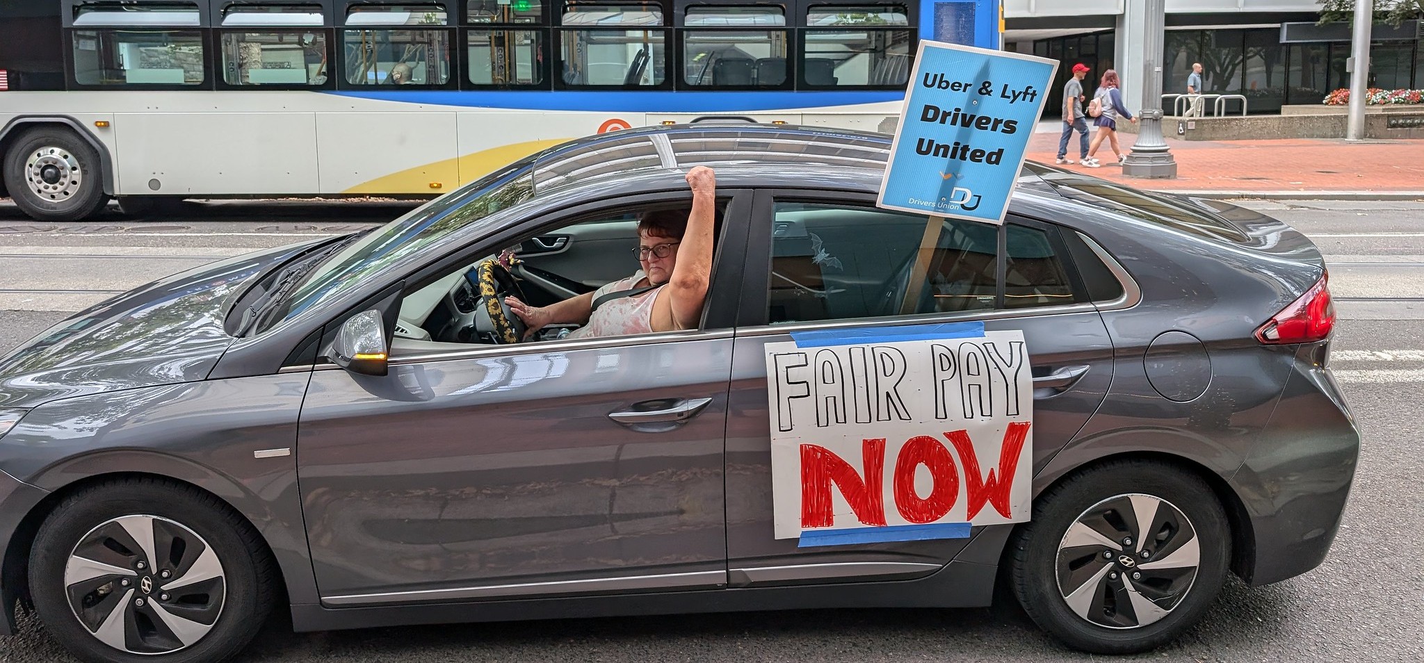 A person driving a car with a picket sign reading "Uber & Lyft Drivers United" and a sign taped to the car reading "Fair Pay Now" smiles and raises their fist.