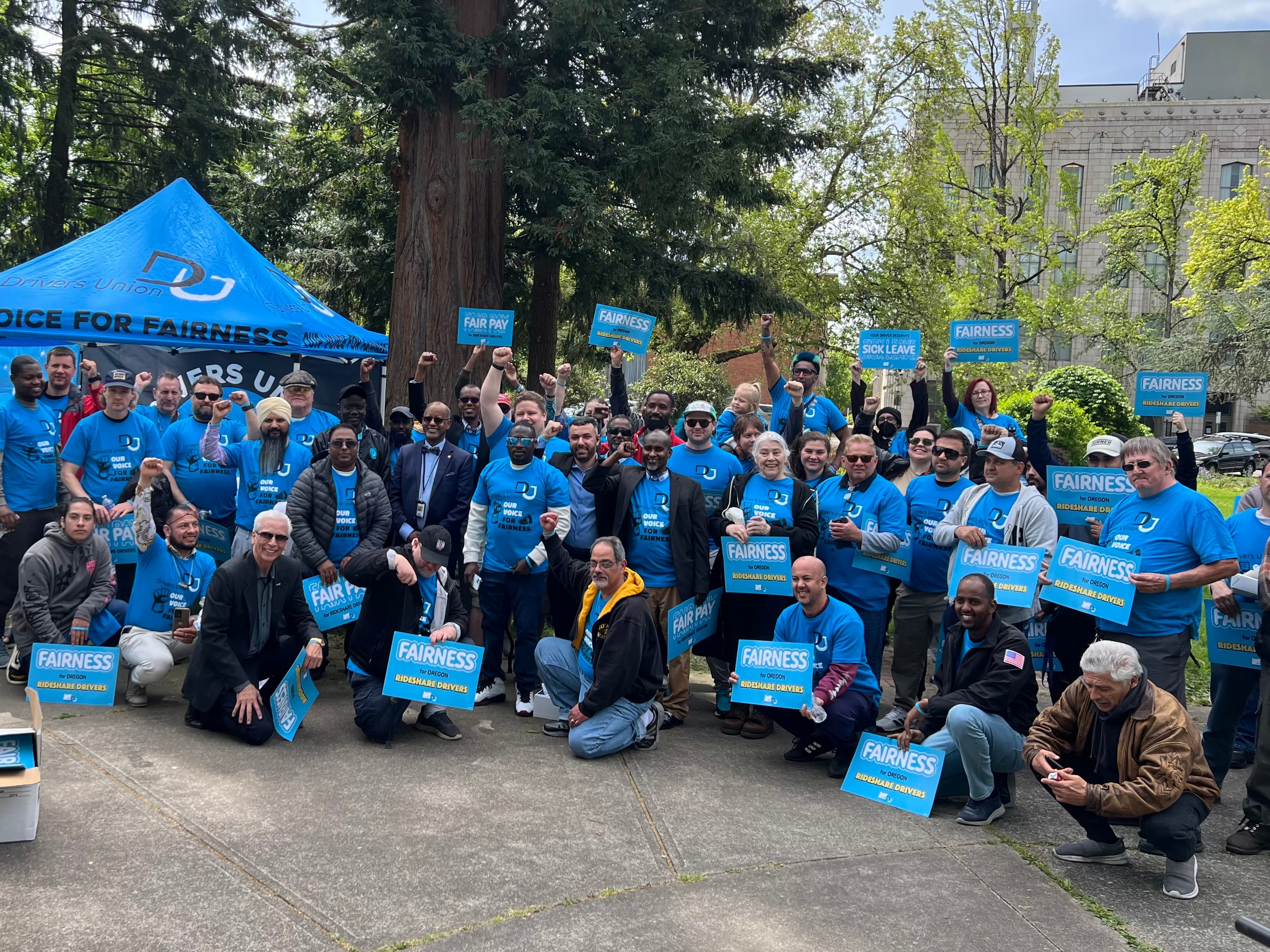 A large crowd of drivers in Drivers Union blue shirts stand for a group photo outside with signs about "Fairness"