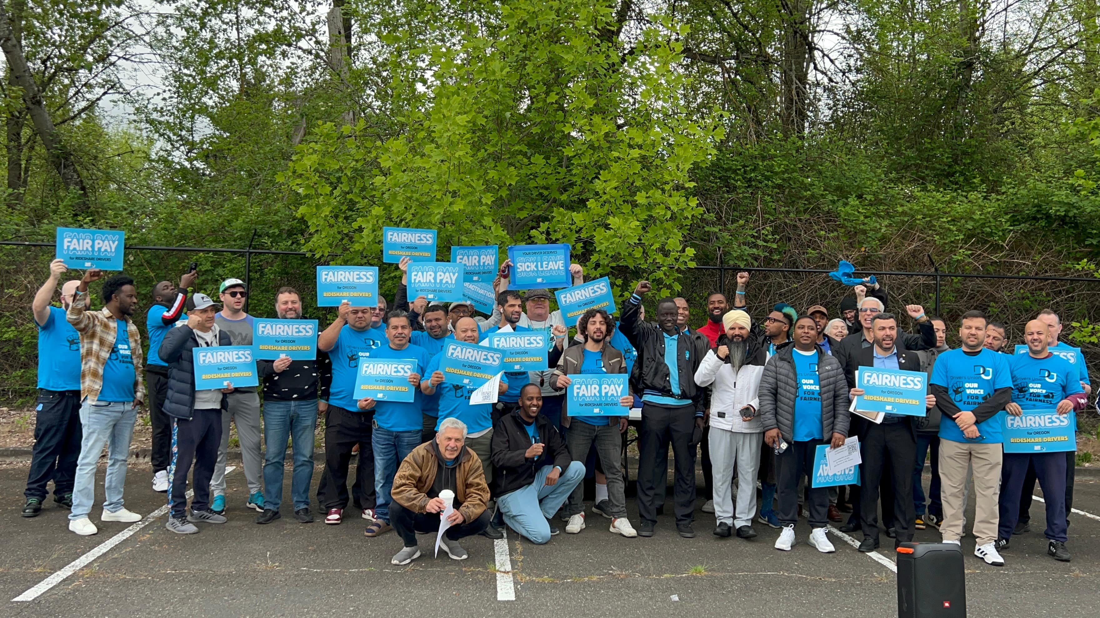A group of drivers in blue Drivers Union shirts holding signs about "FAIRNESS"stands for a group photo on a parking lot