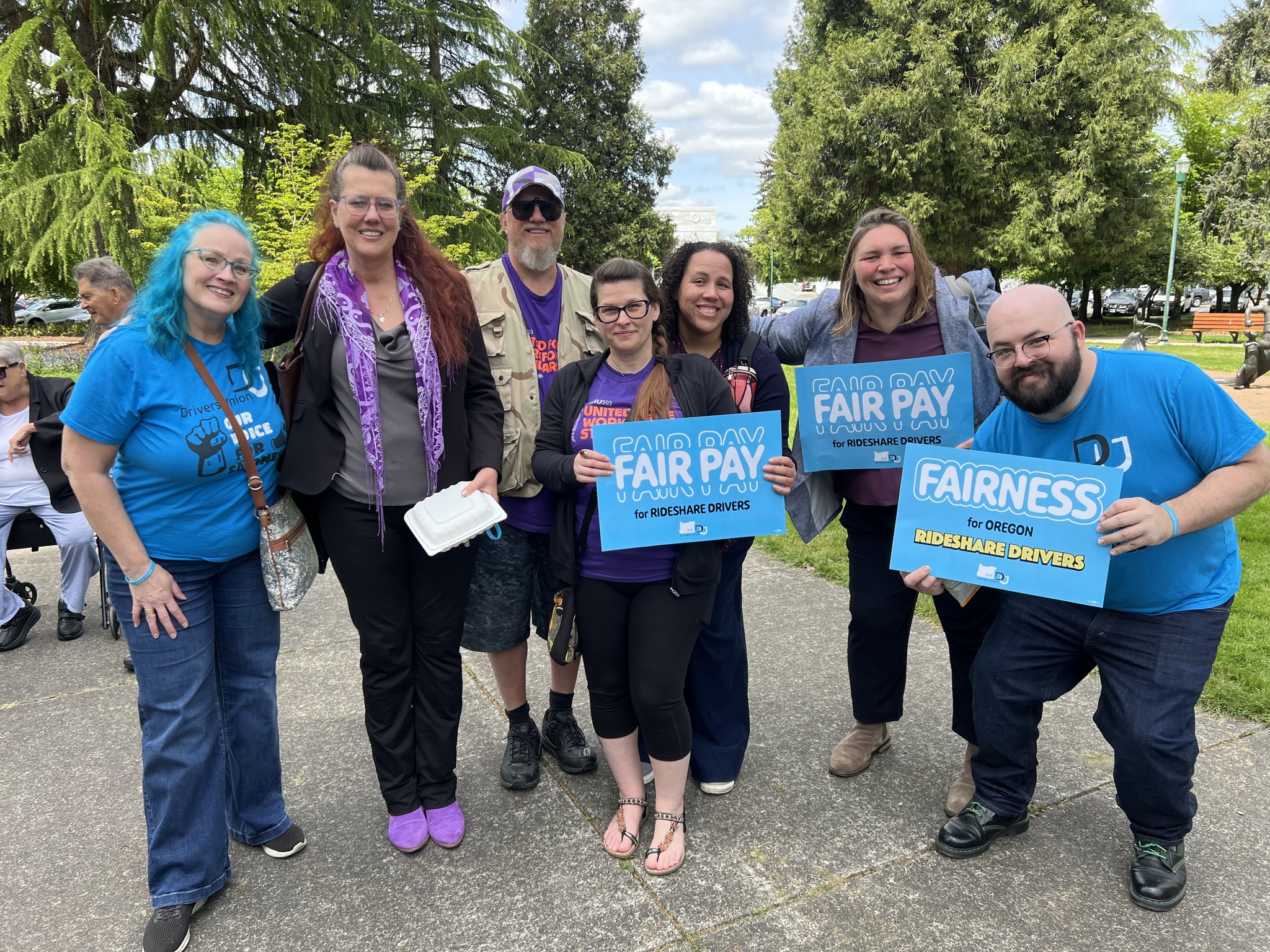 A small group of people, some in purple SEIU shirts and some in blue Drivers Union shirts stand smiling for a group photo outside carrying signs about fairness for drivers.