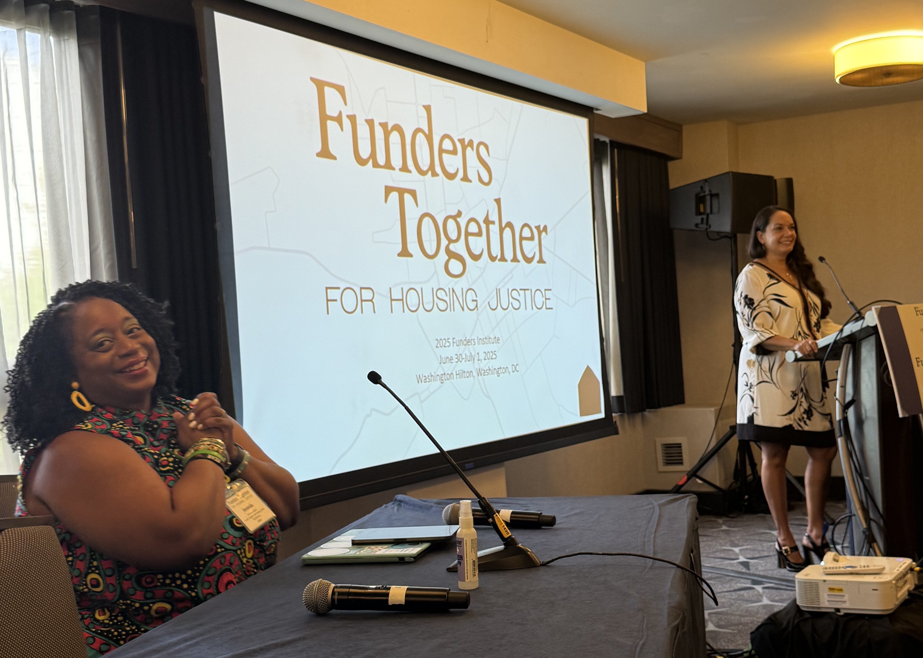 A black woman sits at a table smiling while a Latina woman stands at the podium also smiling