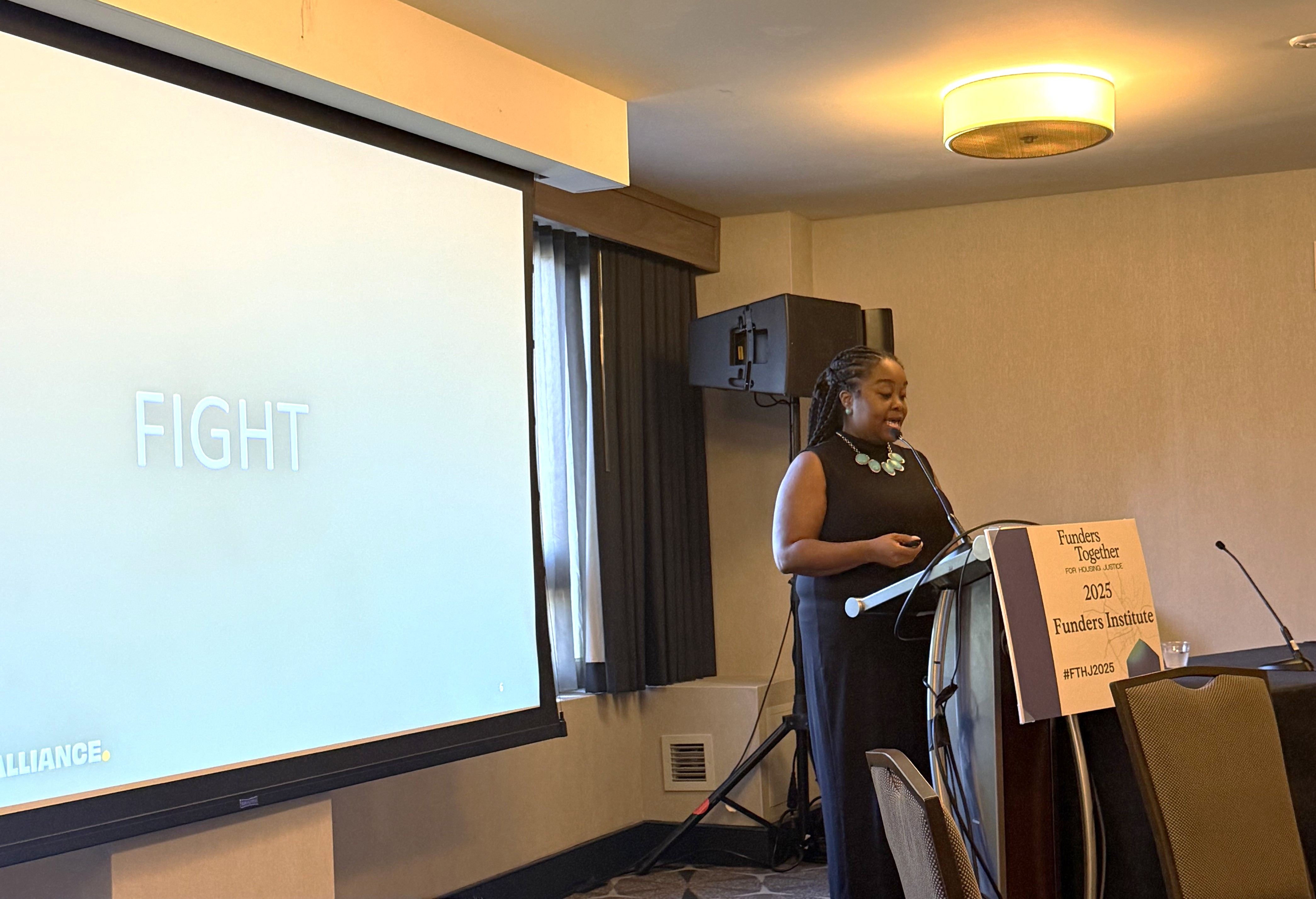 A black woman stands at a podium in front of a screen that reads fight