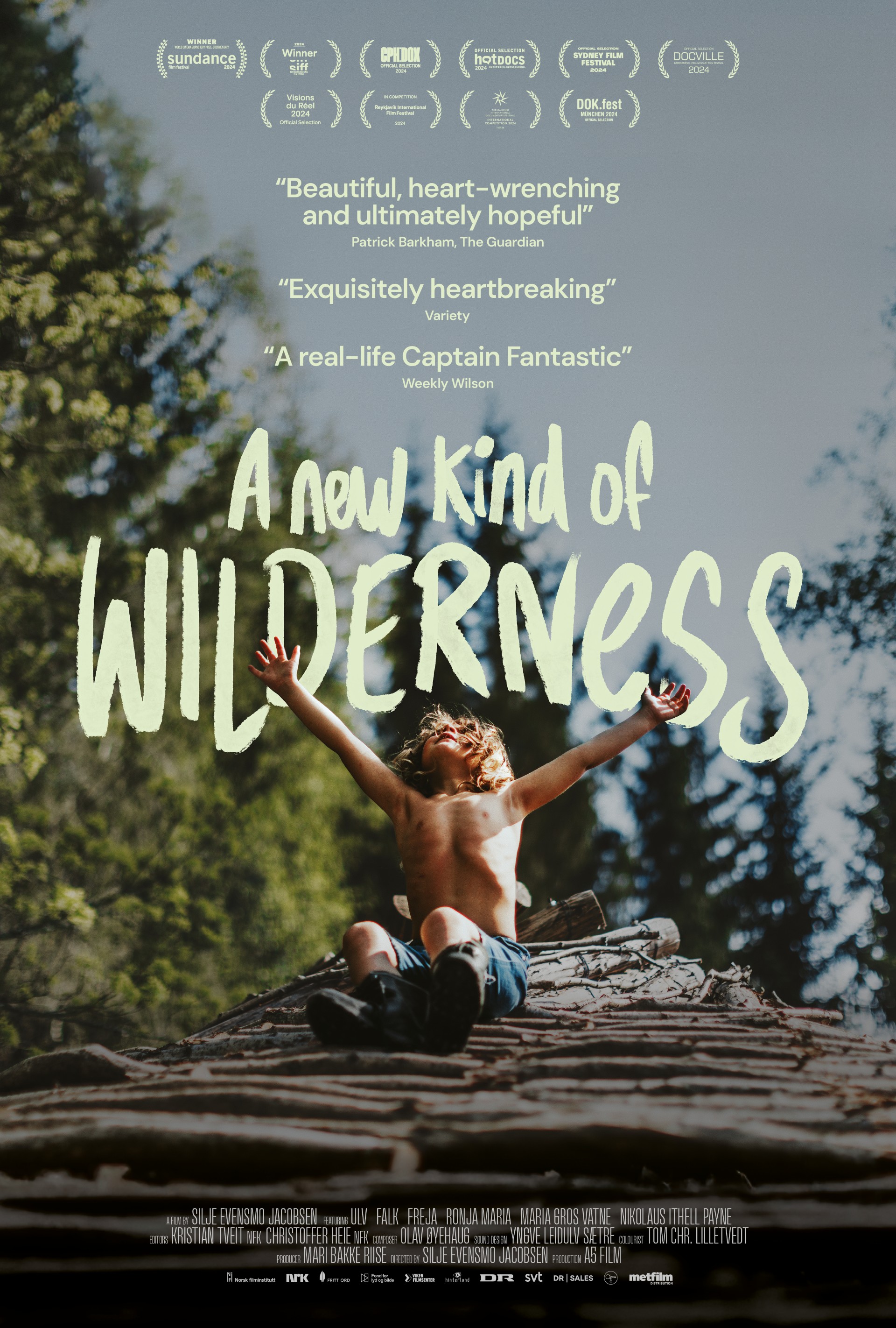 A young boy sits on a rock in the forest, arms thrown wide and head thrown back looking at the sky.