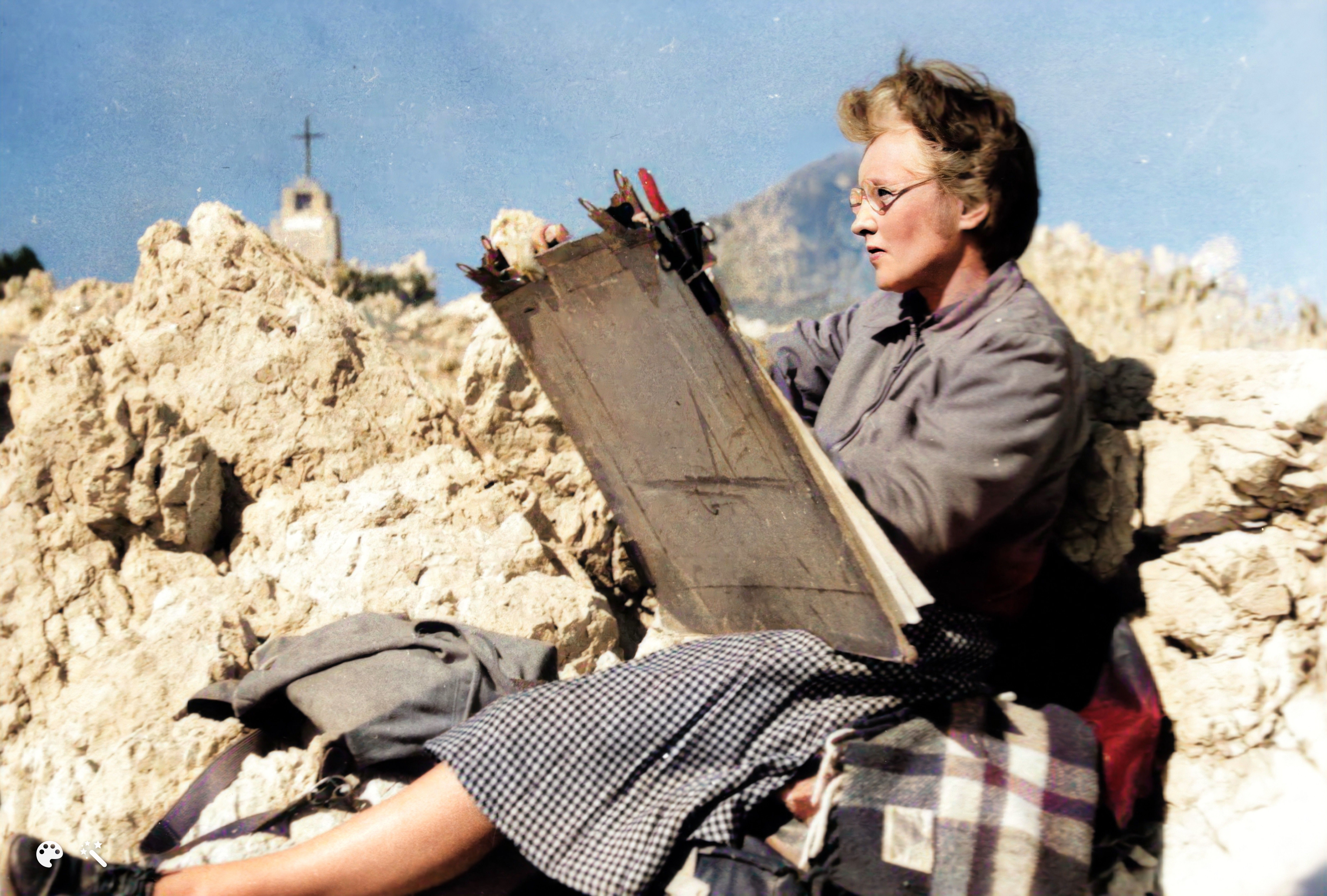 A middle-aged woman with grey hair, a grey wool cardigan, and tartan shawl sits and paints on a large sheet of white paper. She is sitting on a rocky mountaintop, the wind in her hair. 