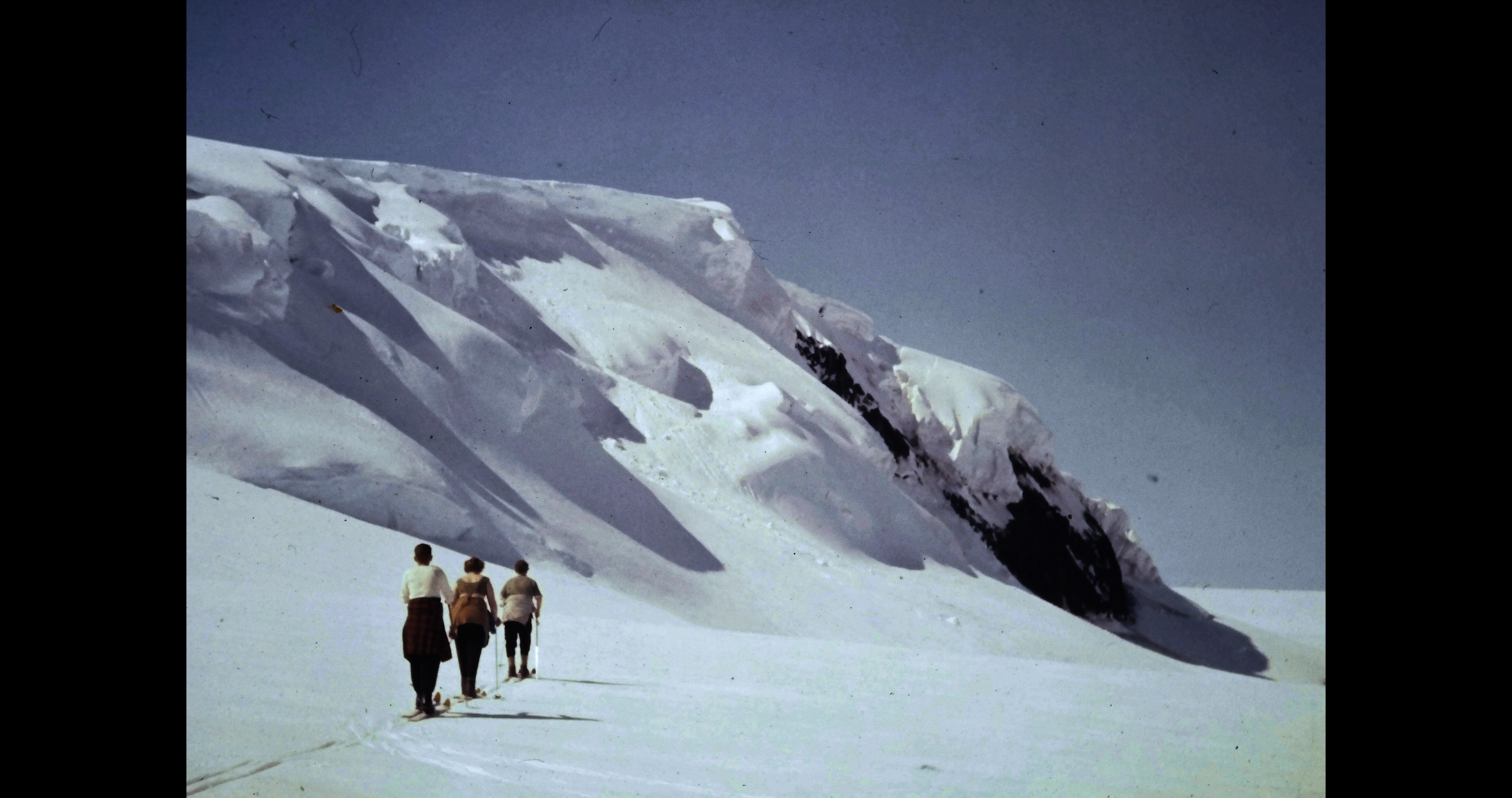 A small group of three cross-country skiers glide across an icy landscape with mountains in the distance.