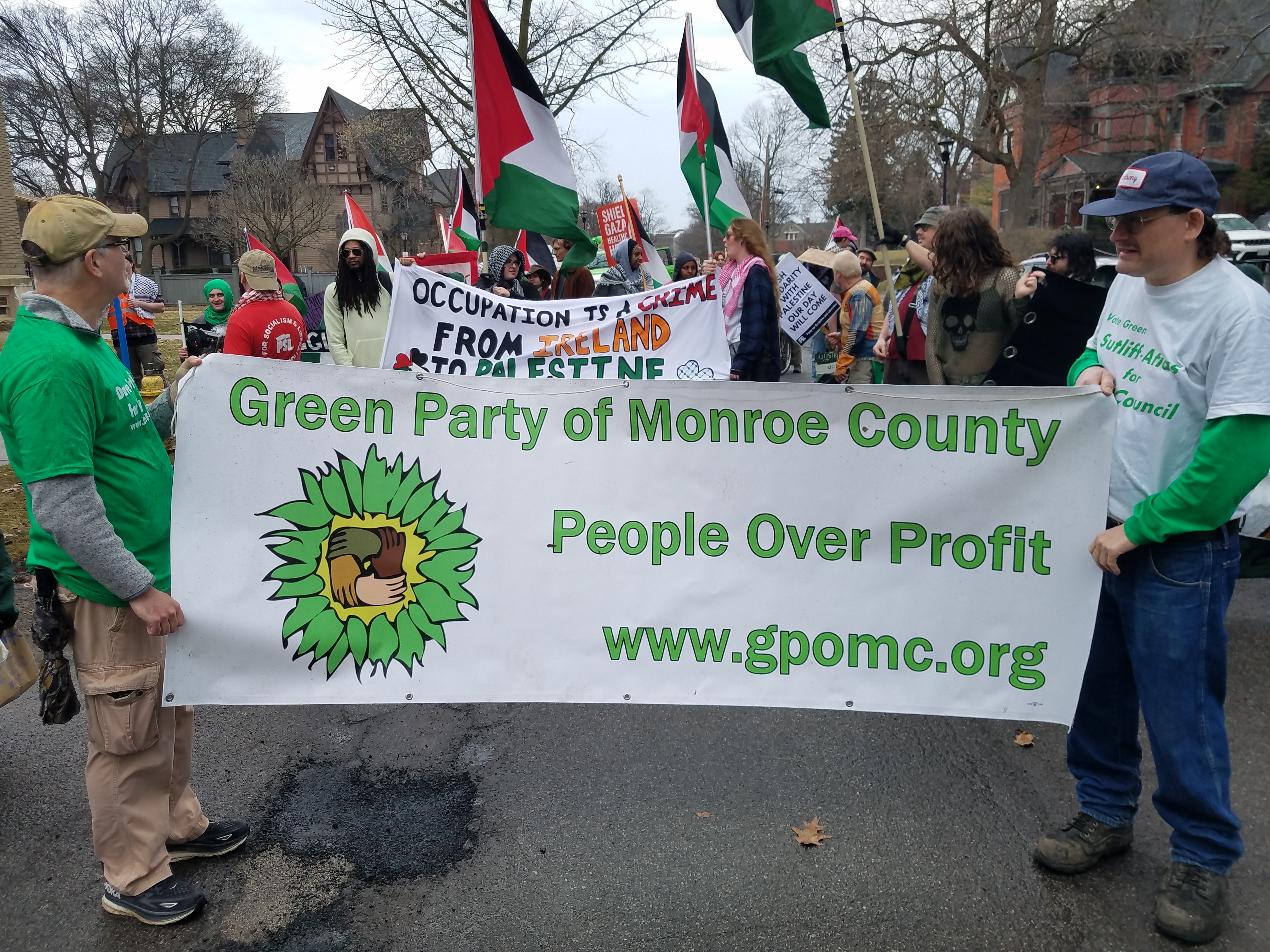 Dozens of people in a parade in downtown Rochester, some holding banners, signs, or flags.