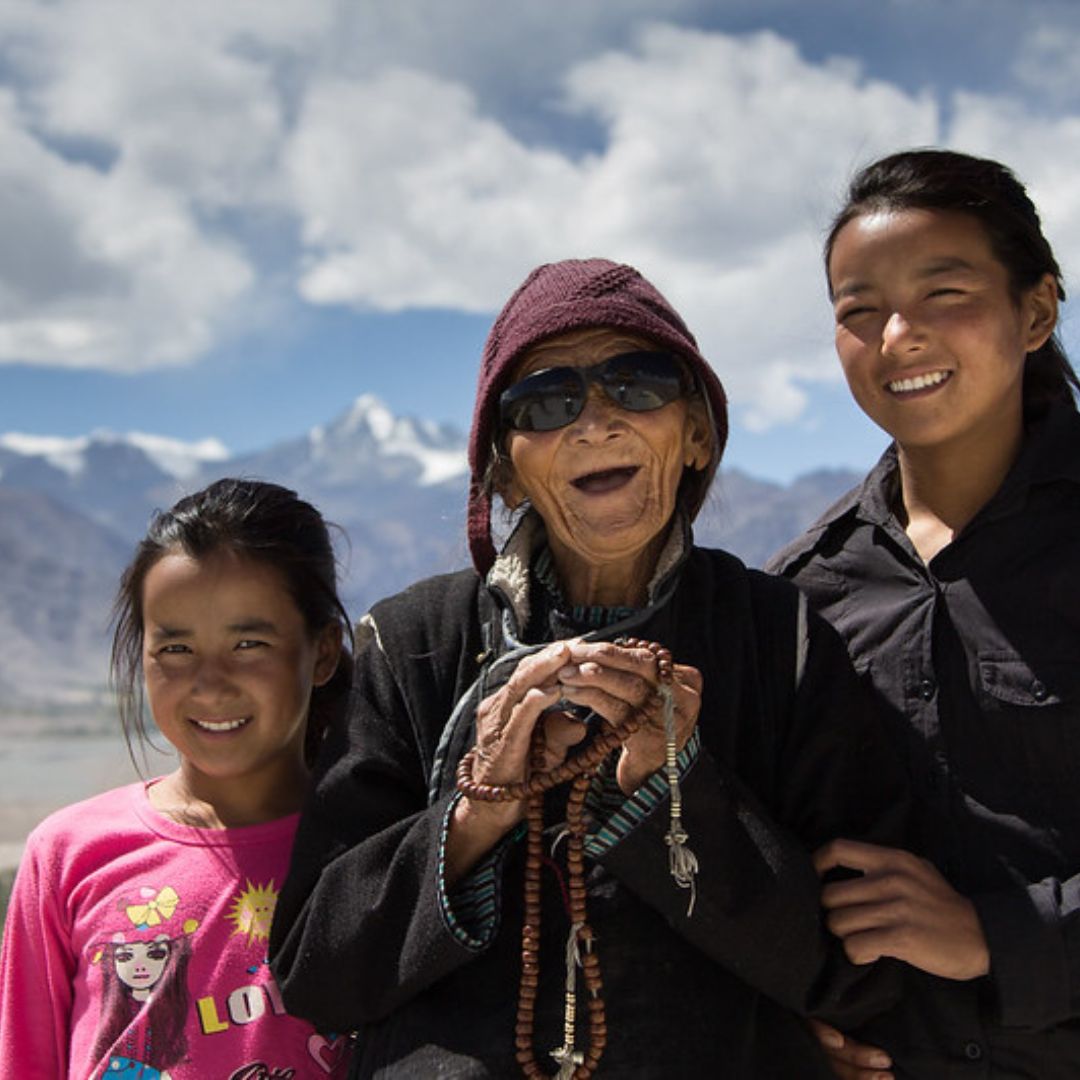Three women stand together smiling with
mountains in the background. The elderly woman in the center wears
glasses.