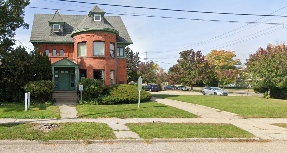 A two-story, red brick Victorian-style home with a green shingledroof and green trim. It has a rounded tower on the front left cornerand a small porch over the main entrance. A concrete sidewalk leads upto the house, and a paved driveway runs along the right side of theyard, where several cars are parked. The lawn is green with a fewtrees, including one with changing autumn leaves. The sky is clearwith power lines visible in the distance.