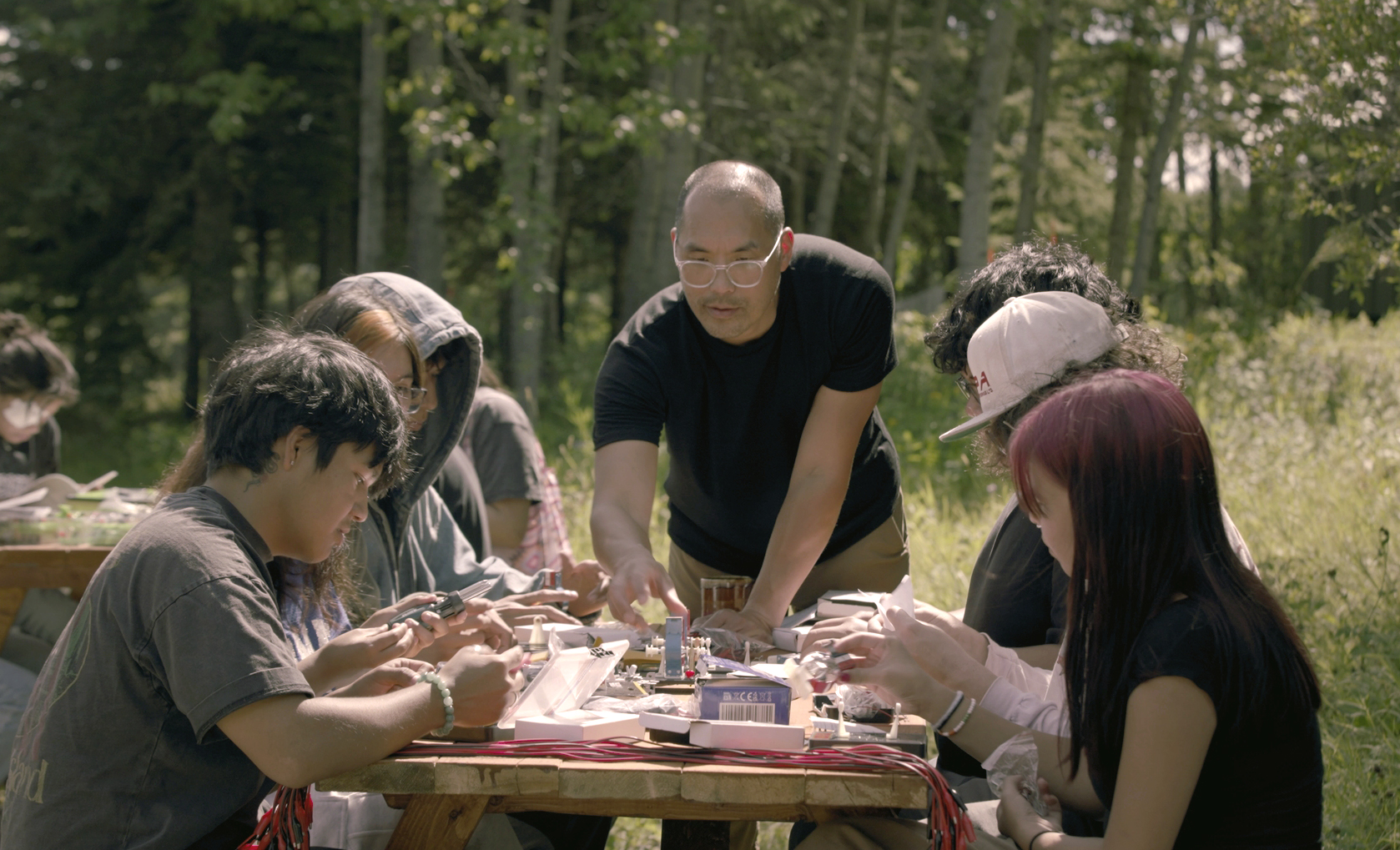 Youth sit at a picnic table while and instructor demonstrates how to put together simplified solar panel wirings.