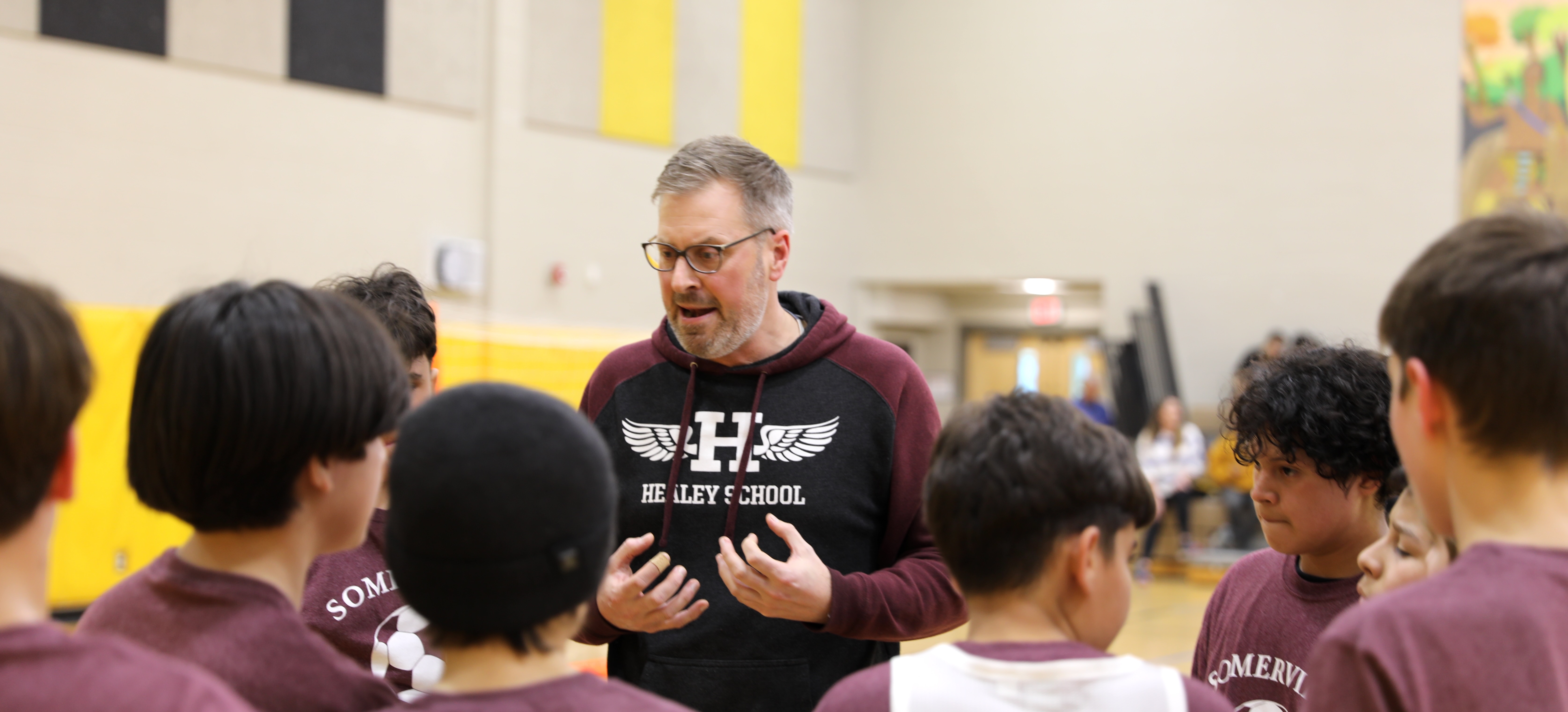 Jake, wearing a red Healey School hoodie, talks with a group of youth in a gymnasium