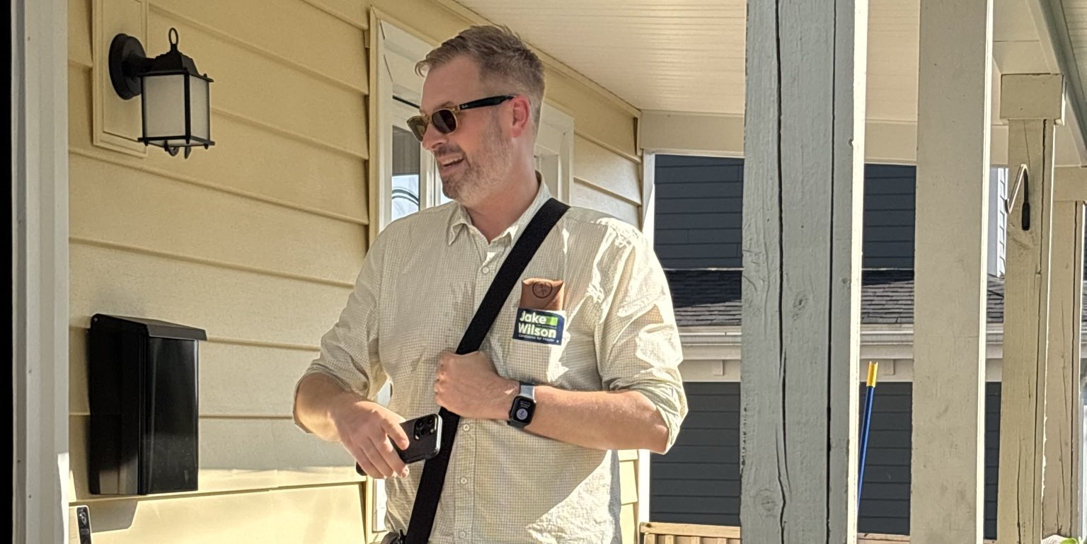 Jake, wearing sunglasses and a button down shirt with the sleeves rolled up, holds a conversation with someone on their porch