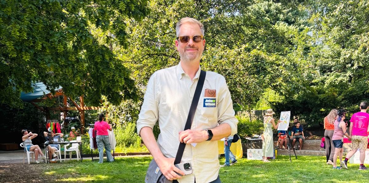 Jake in a park wearing a satchel, sunglasses, and a button down shirt on a sunny day.