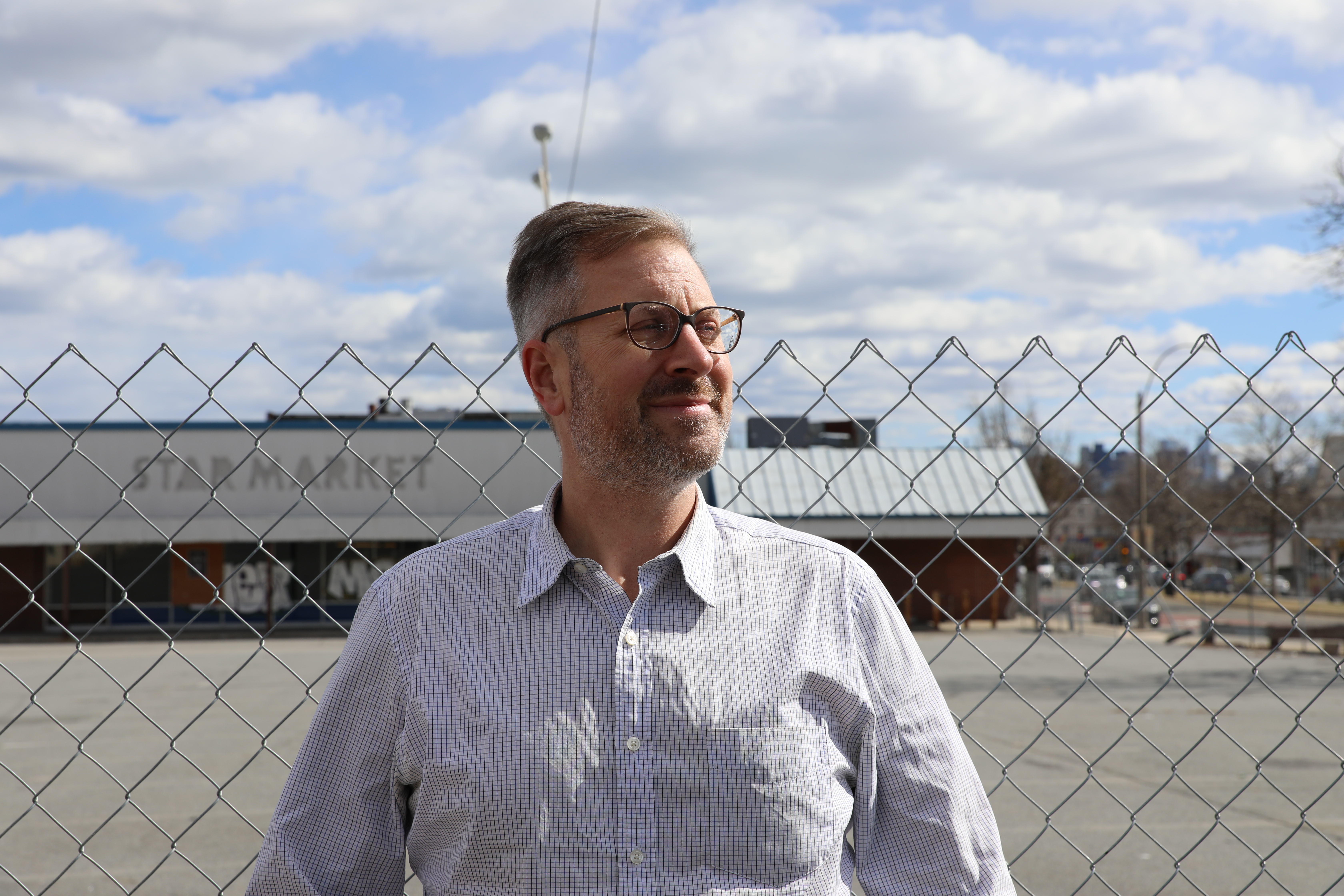 Jake posing in front of a chain-link fence that surrounds an abandoned grocery store lot