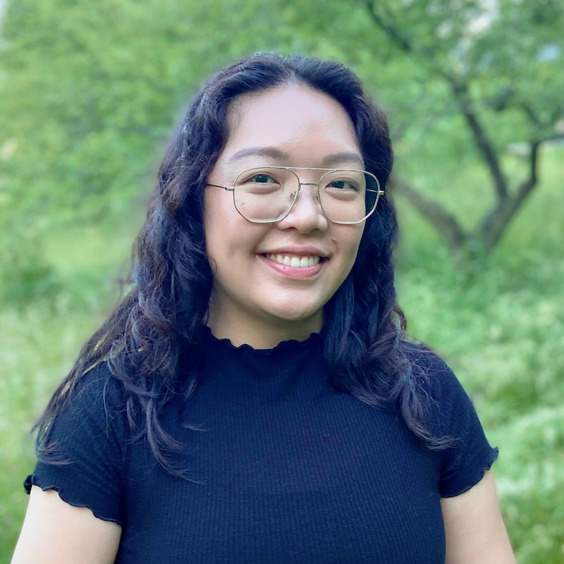 Angela, an Asian-American woman with thick, wavy dark brown hair, wearing glasses with metallic rims, smiles at the camera, showing her teeth. The background is filled with green foliage in the distance.