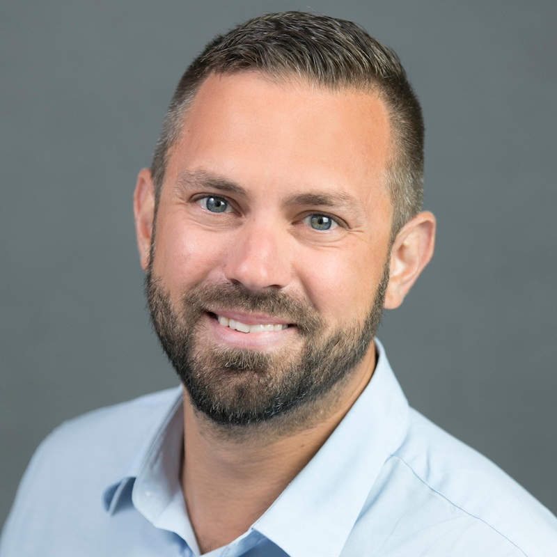 Chris, a white man with grey-blue eyes, brown hair, and a short beard, is standing at a slight angle but is looking at the viewer. He is smiling with his teeth showing; he is wearing a light-blue collared shirt, and there is a grey flat background.