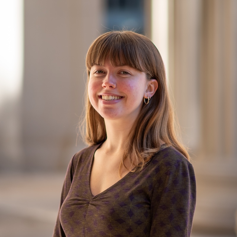 Sophie, a young white woman with straight light brown hair and bangs, stands at a slight angle and smiles at the viewer, showing her teeth. She is wearing a brown v-neck shirt, and the background appears to be out of focus pillars.