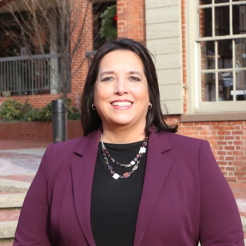 a photo of lieutenant governor candidate kim driscoll, a white woman with shoulder-length straight brown hair, wearing a purple blazer, a black shirt, and a colorful necklace, smiling at the camera with her teeth showing.