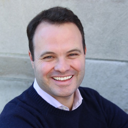 eric lesser, a candidate for lieutenant governor, smiles at the camera with his mouth open and teeth showing. he is a white man with dark brown hair and a high, receding hairline, and he is wearing a white collared shirt under a dark sweater