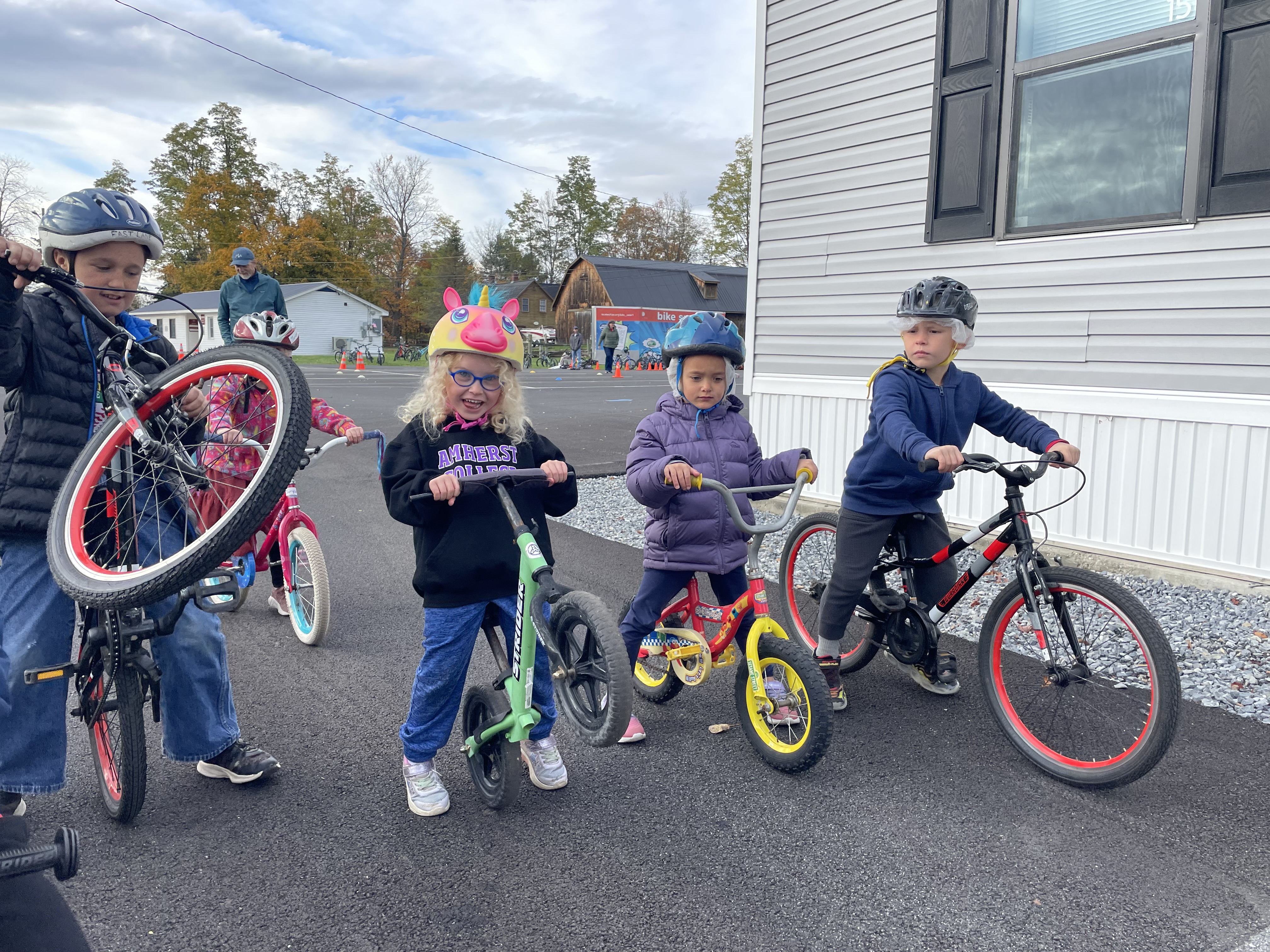 Four children stand over bicycles. They are lifting up the front wheels of their bicycles. All of them are smiling and wearing helmets!
