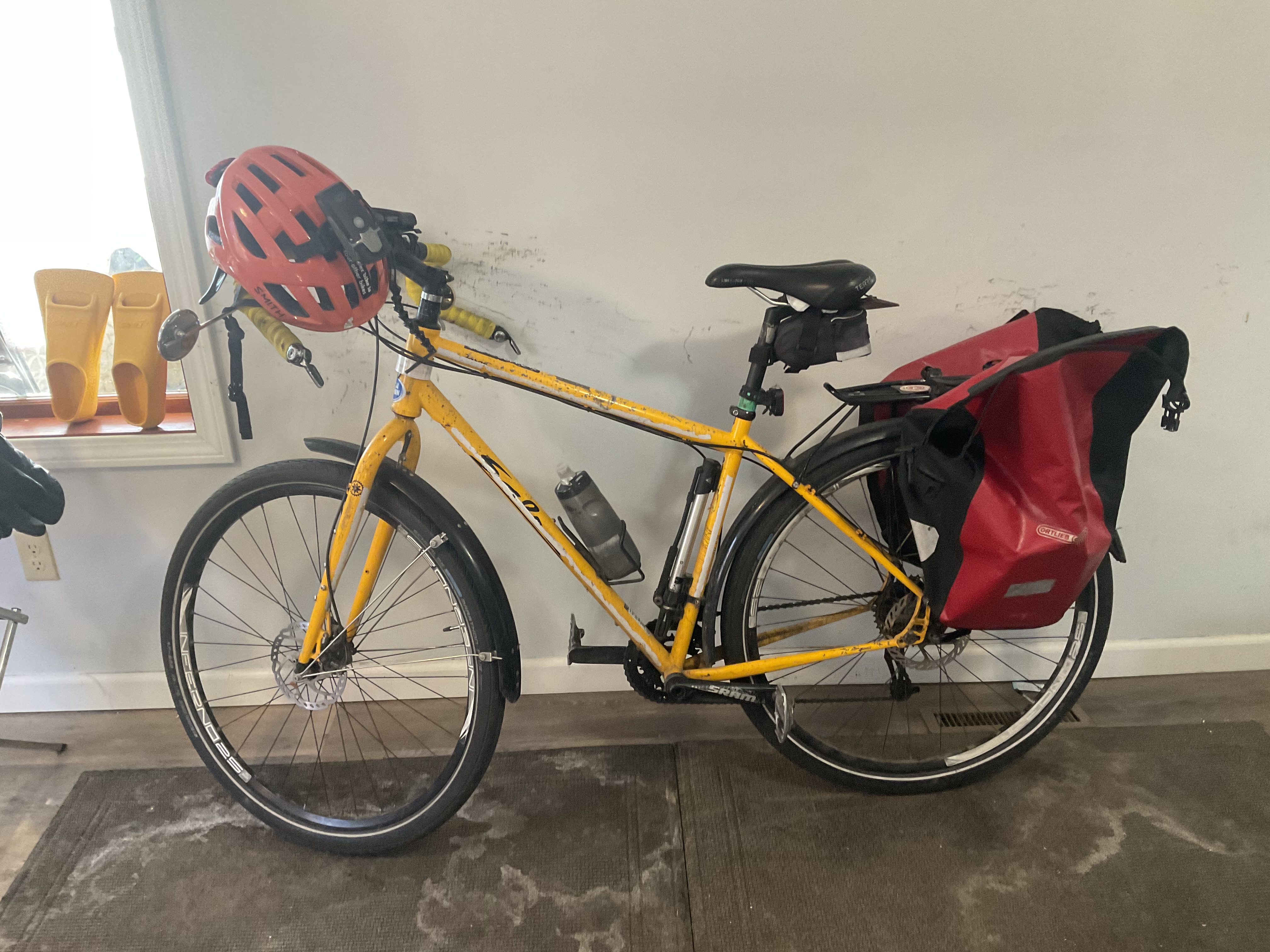 Peter's bike has a rack on the back with two pannier bags installed. his helmet is hanging off the handlebars. on his helmet are two lights and a mirror.