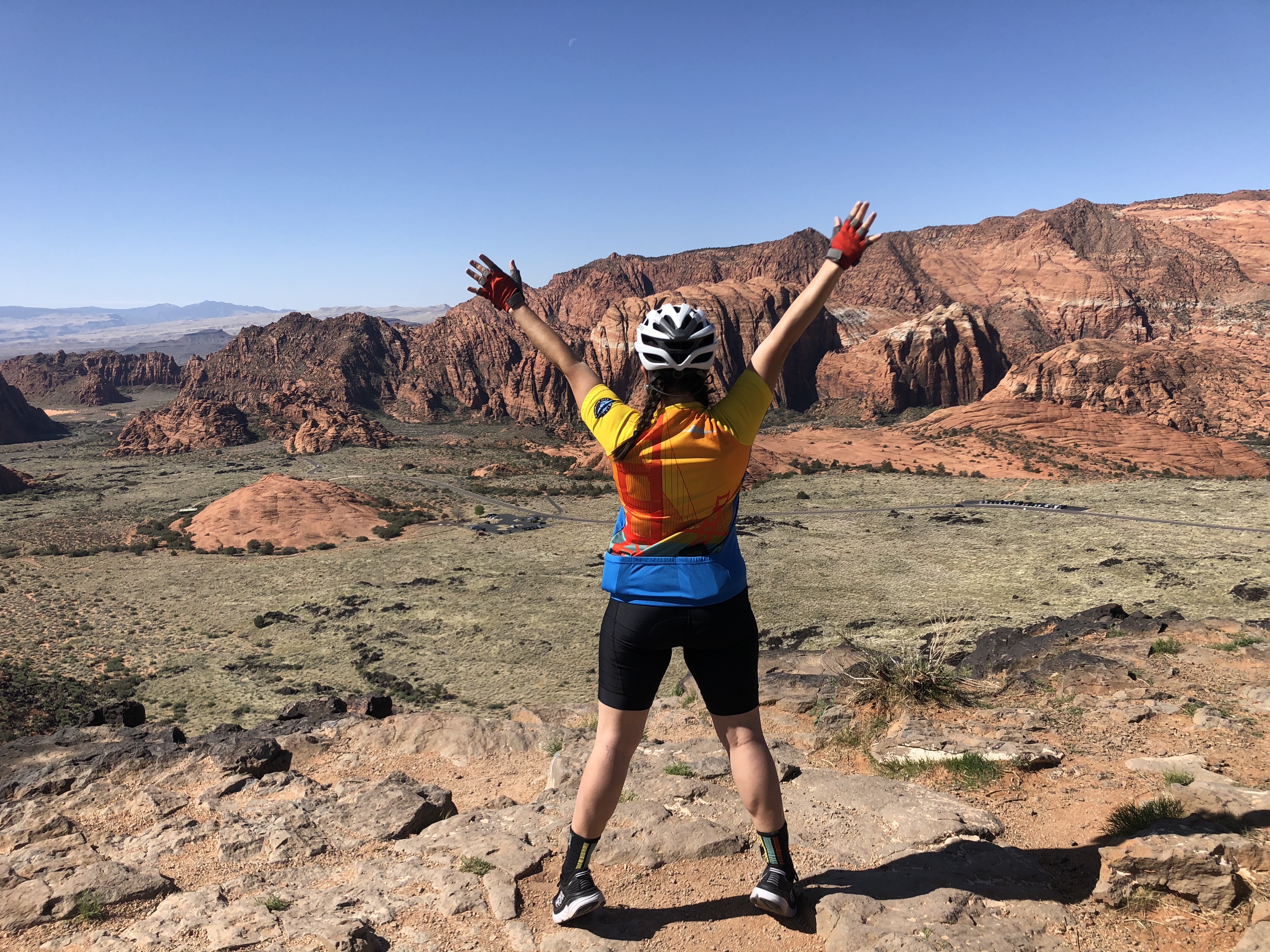A woman in cycling kit stands at the edge of a canyon in Utah, with her arms stretched out above her head in celebration