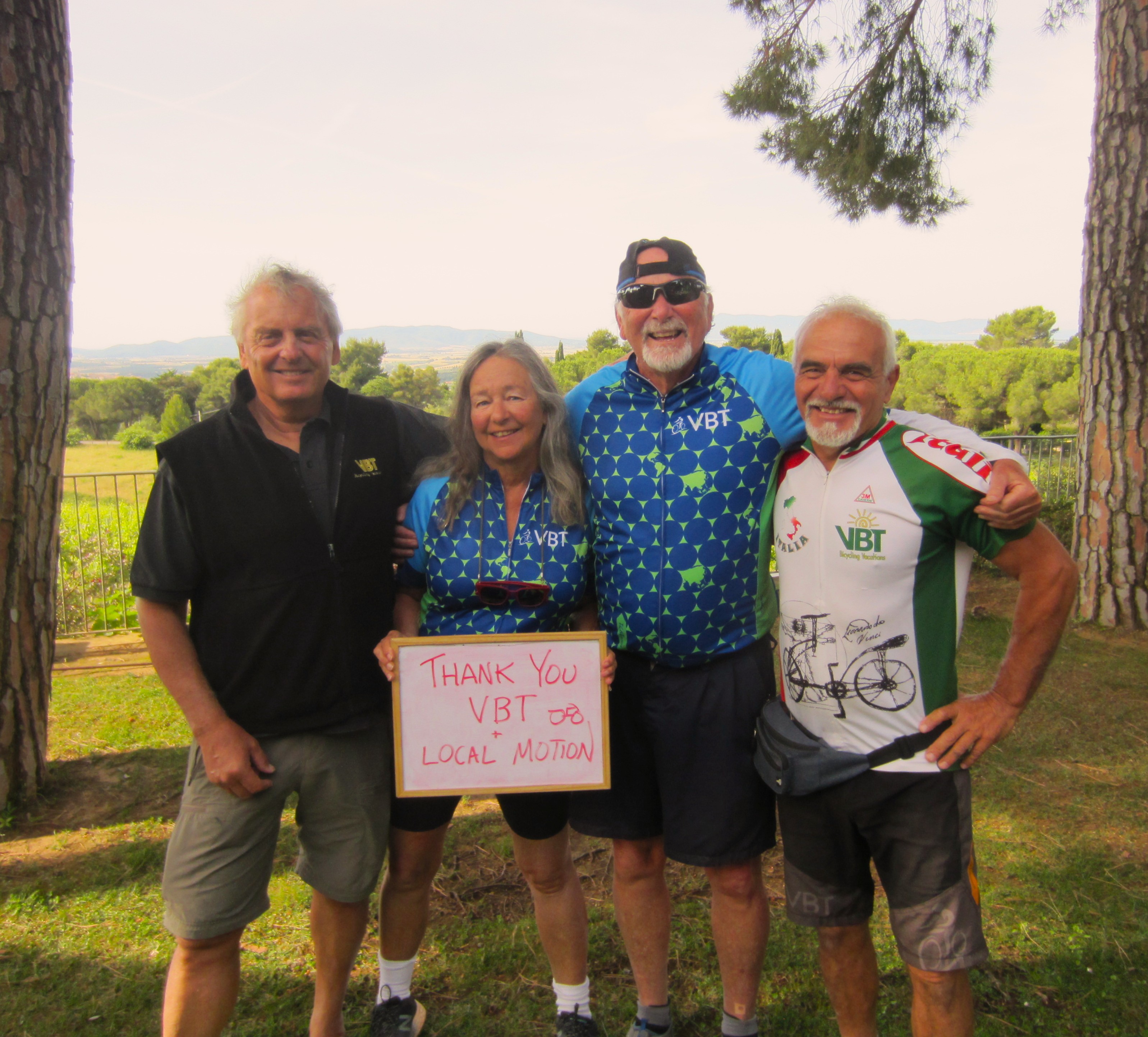 Four people stand together and smile in front of an Italian villa in Tuscany. A woman in the group holds a sign that reads 'thanks local motion and VBT'