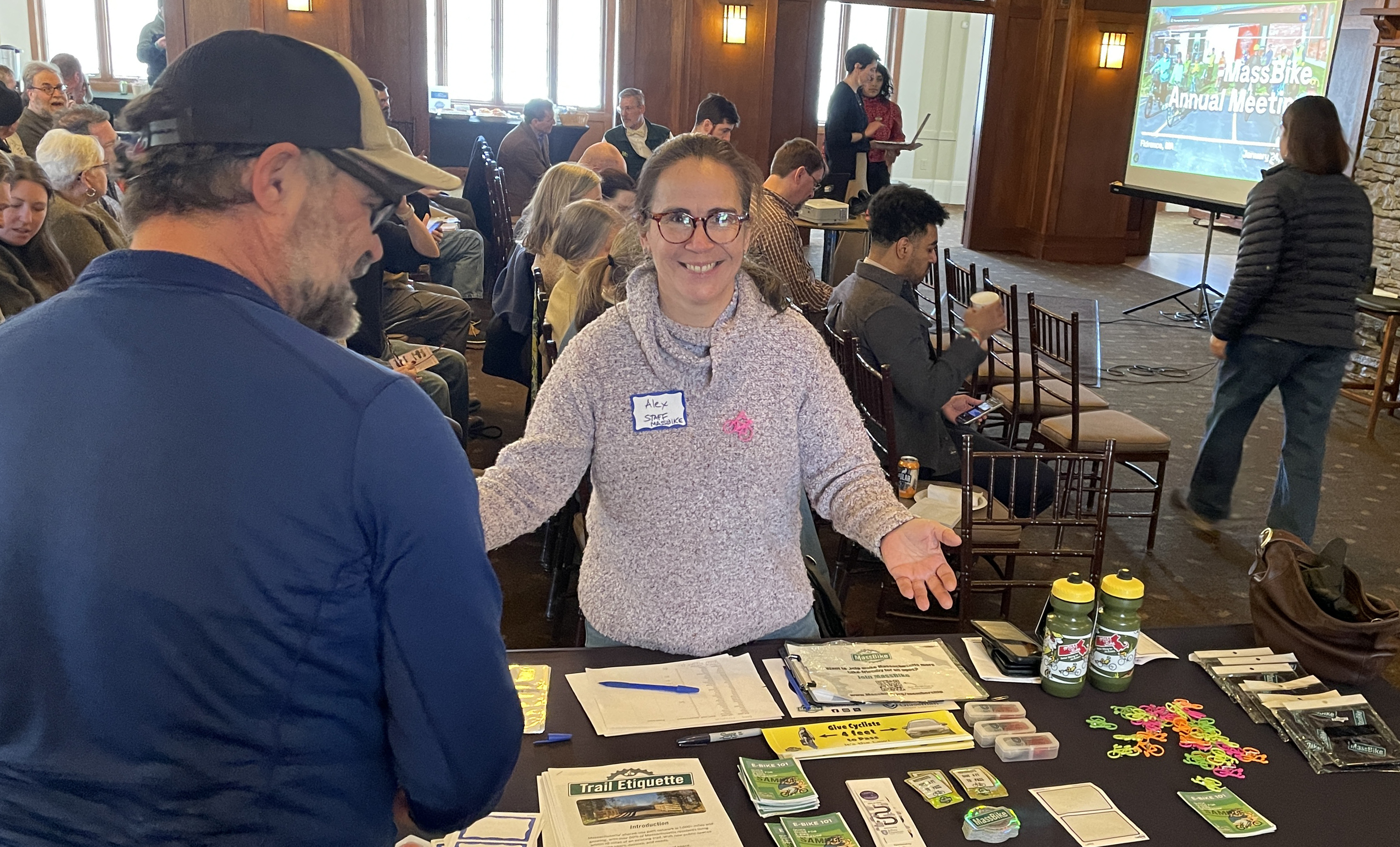 Alex, a light-skinned hispanic woman, stands behind a desk with registration materials on it. she is wearing a light purple sweater and a nametag, and smiling at the camera with her arms held open. in the open room behind her, people are sitting in rows of chairs in preparation for the presentation. there is a light-skinned man in front of the table with his back to us.