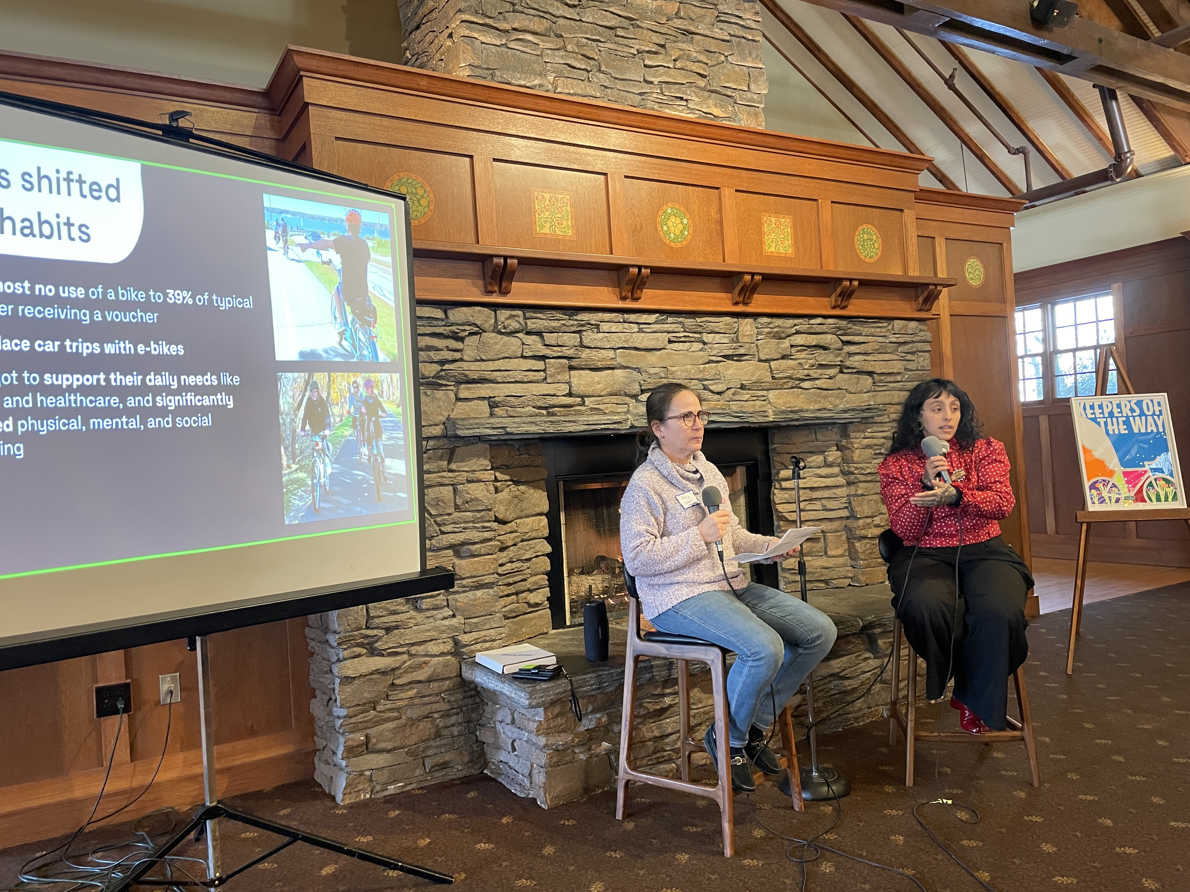 Alex and Sara, two light-skinned latina women, sit in bar-height stools facing the audience (off camera right). they are both holding microphones, and sara is speaking into hers as she gestures with her hand. on the left side of the picture is a portion of a projector screen with a slide displayed on it. on the right is an easel with a "keepers of the way" poster on it. alex and sara are sitting in front of a stone fireplace with a fire in it.