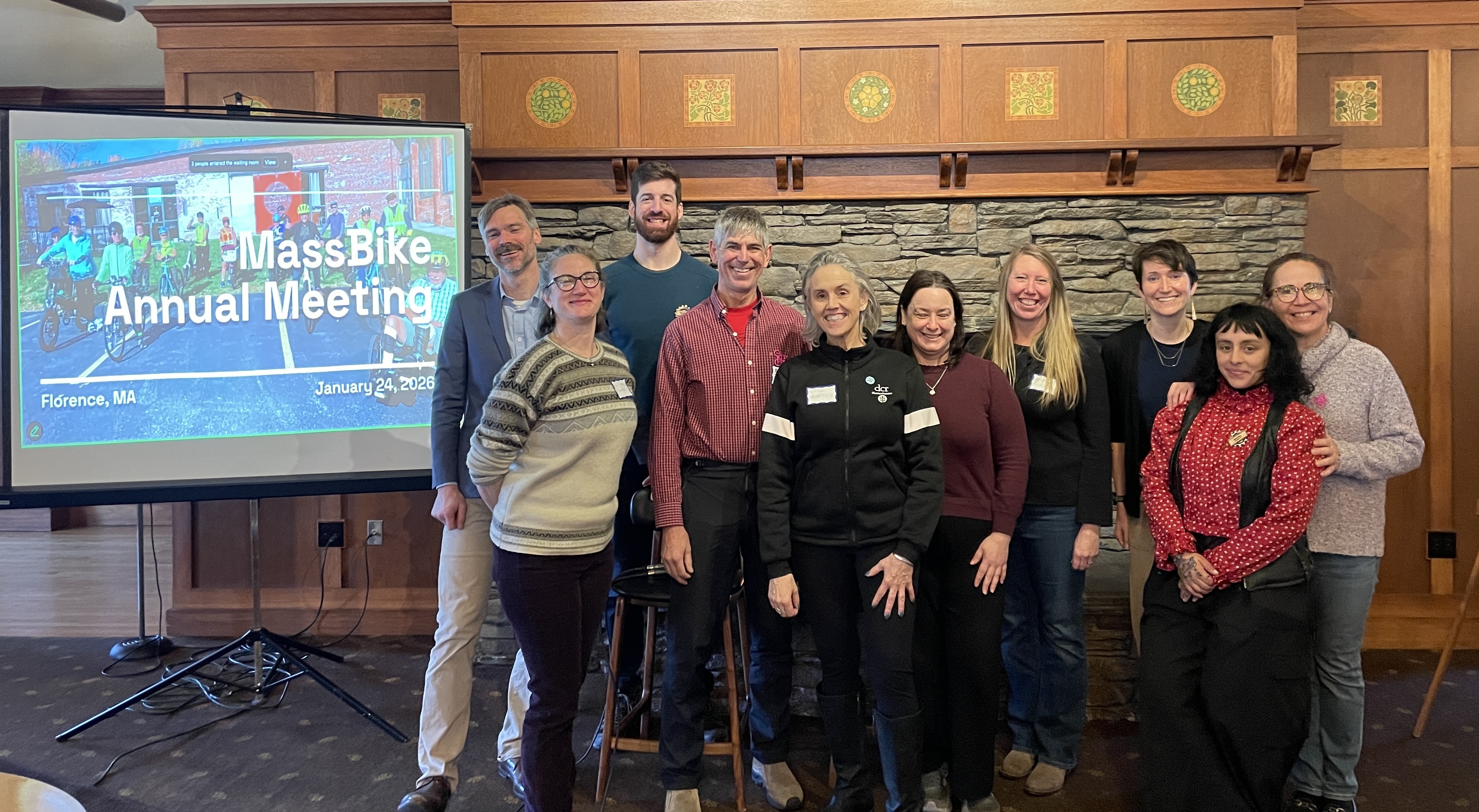 the massbike team (+ others) stand in front of a stone fireplace for a group picture. on camera left is a projector screen showing a slide that says "massbike annual meeting". the ten people are all looking at the camera and smiling