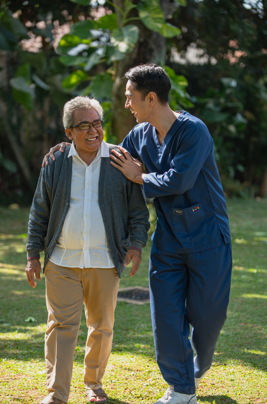 An aged care worker walking outside with older person. The worker has a he/him pronoun pin and an 'ally' pin on his pocket.