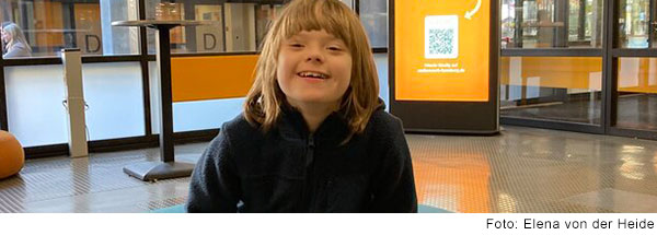 Ein Junge mit mittellangen braunen Haaren und Trisomie 21 sitzt auf einer Bank und lacht. 