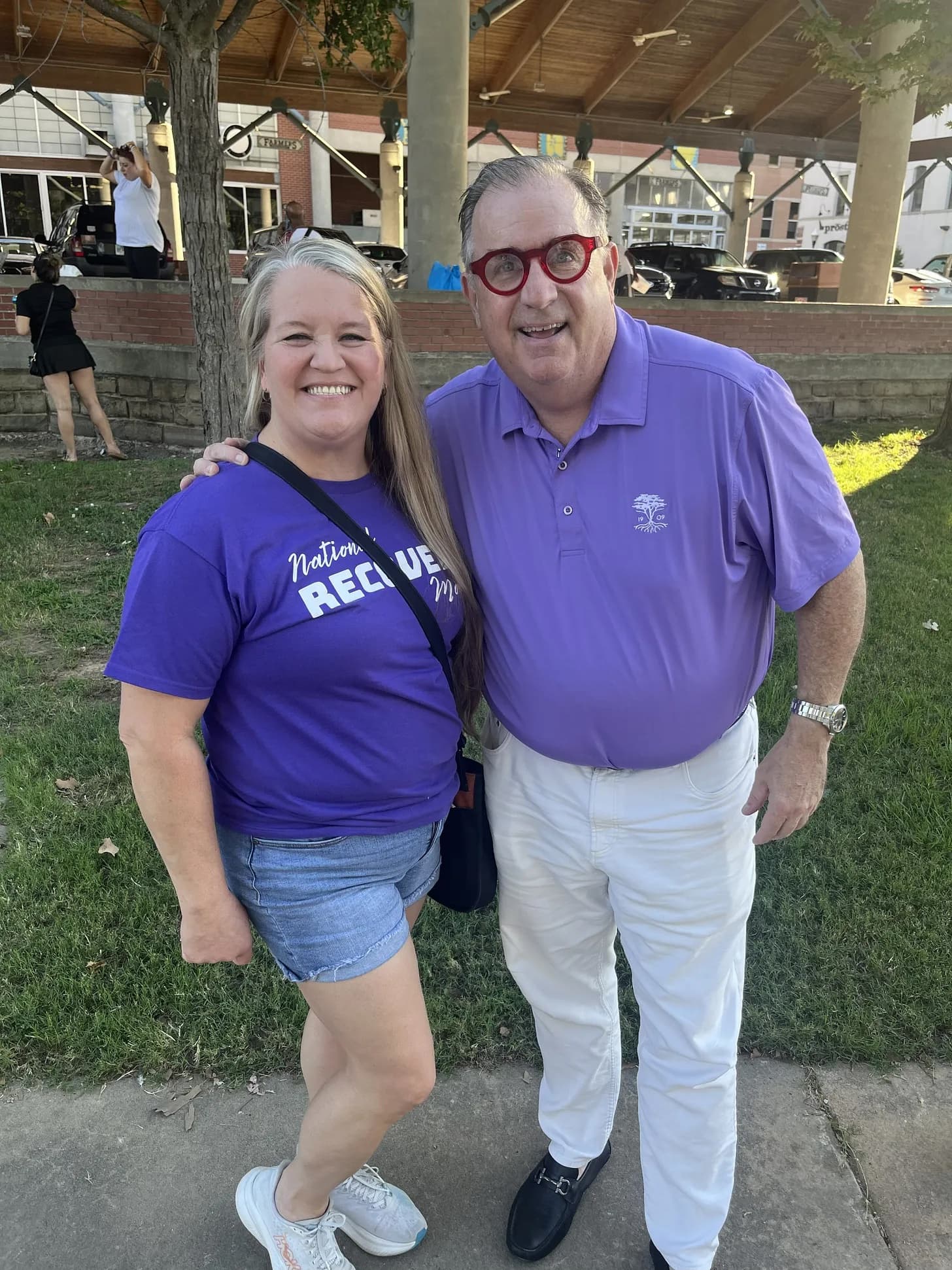 Tom Farley smiling outdoors beside a woman, both facing the camera in daylight with trees and buildings visible in the background.