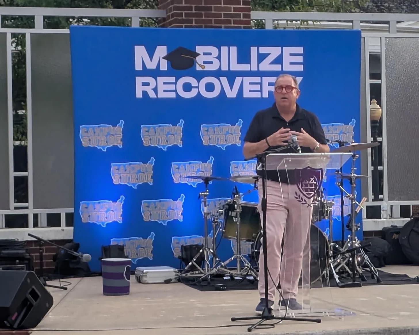 Tom Farley speaking at a podium in front of a blue ‘Mobilize Recovery – Campus Surge’ backdrop.