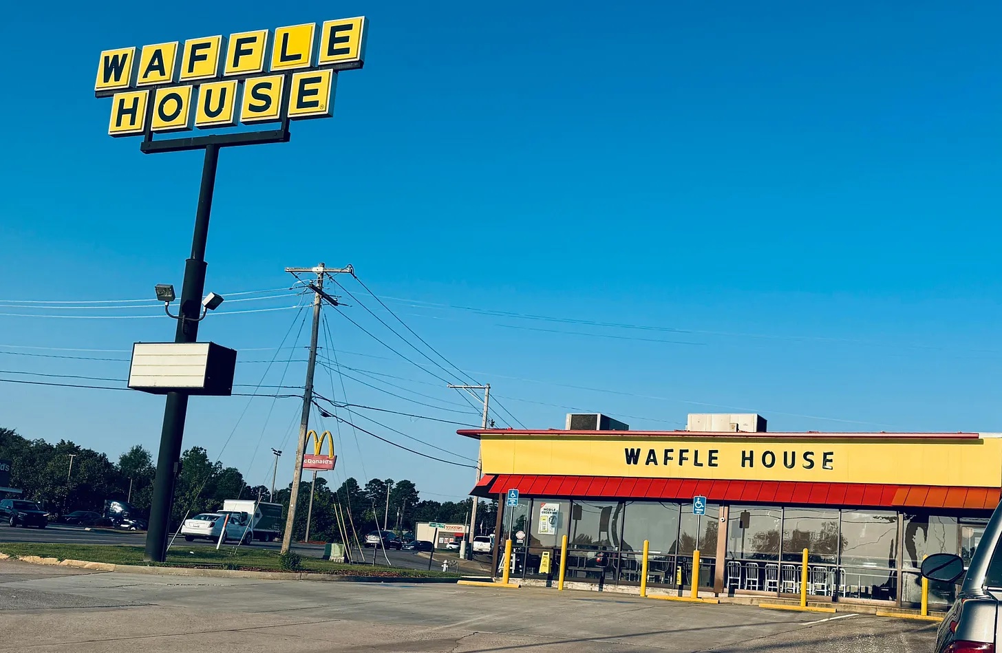 A Waffle House restaurant and roadside sign under a clear blue sky, with parking lot and power lines in view.