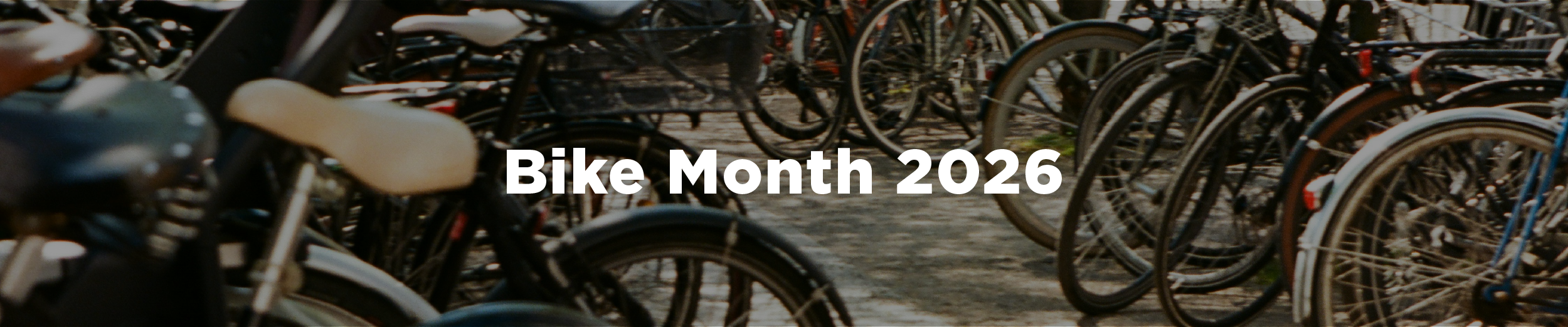 A group of bicycles locked to bike racks on a quiet urban street with the overlaid text "Bike Month 2026"