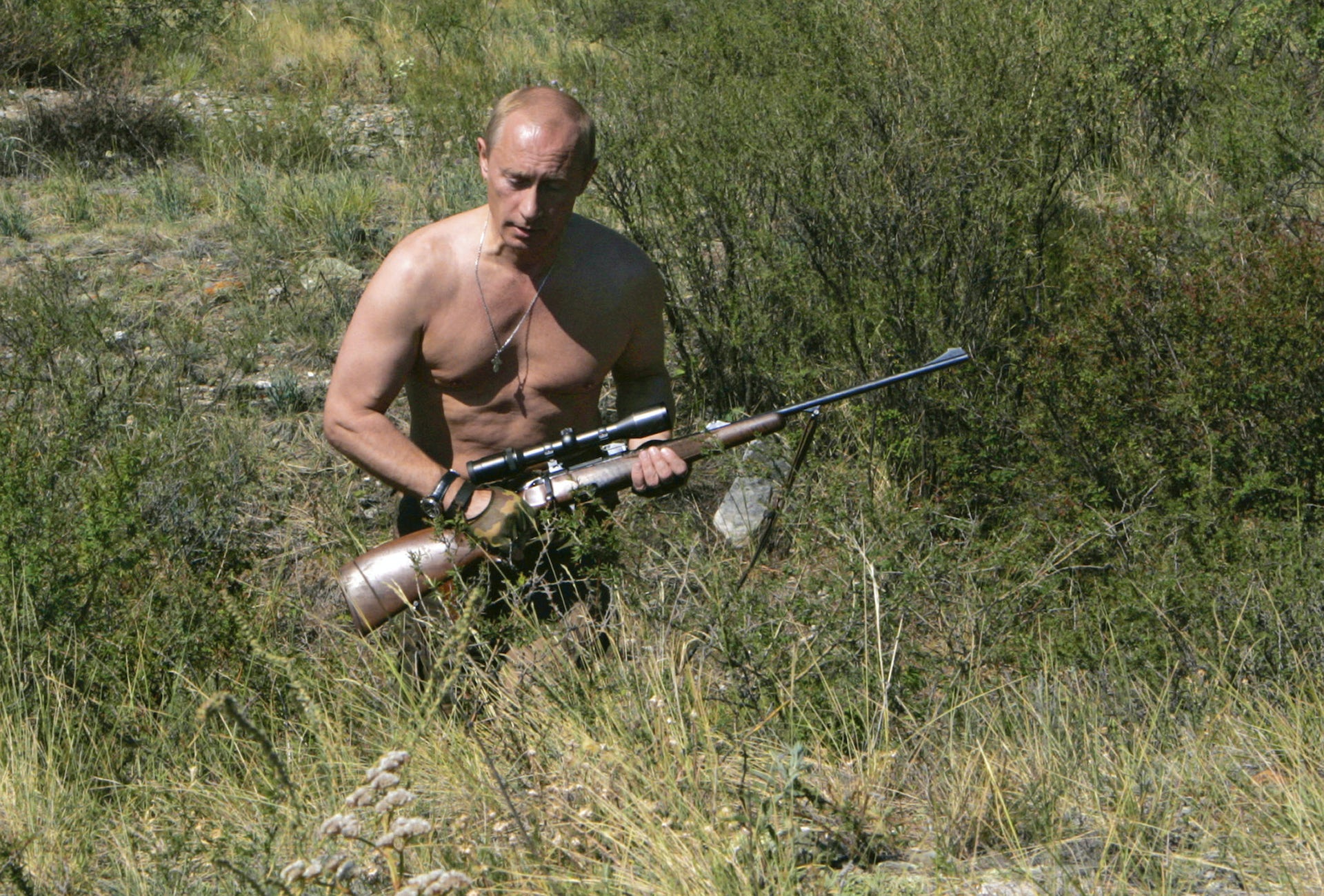 A shirtless balding man walks through a field clutching a rifle.
