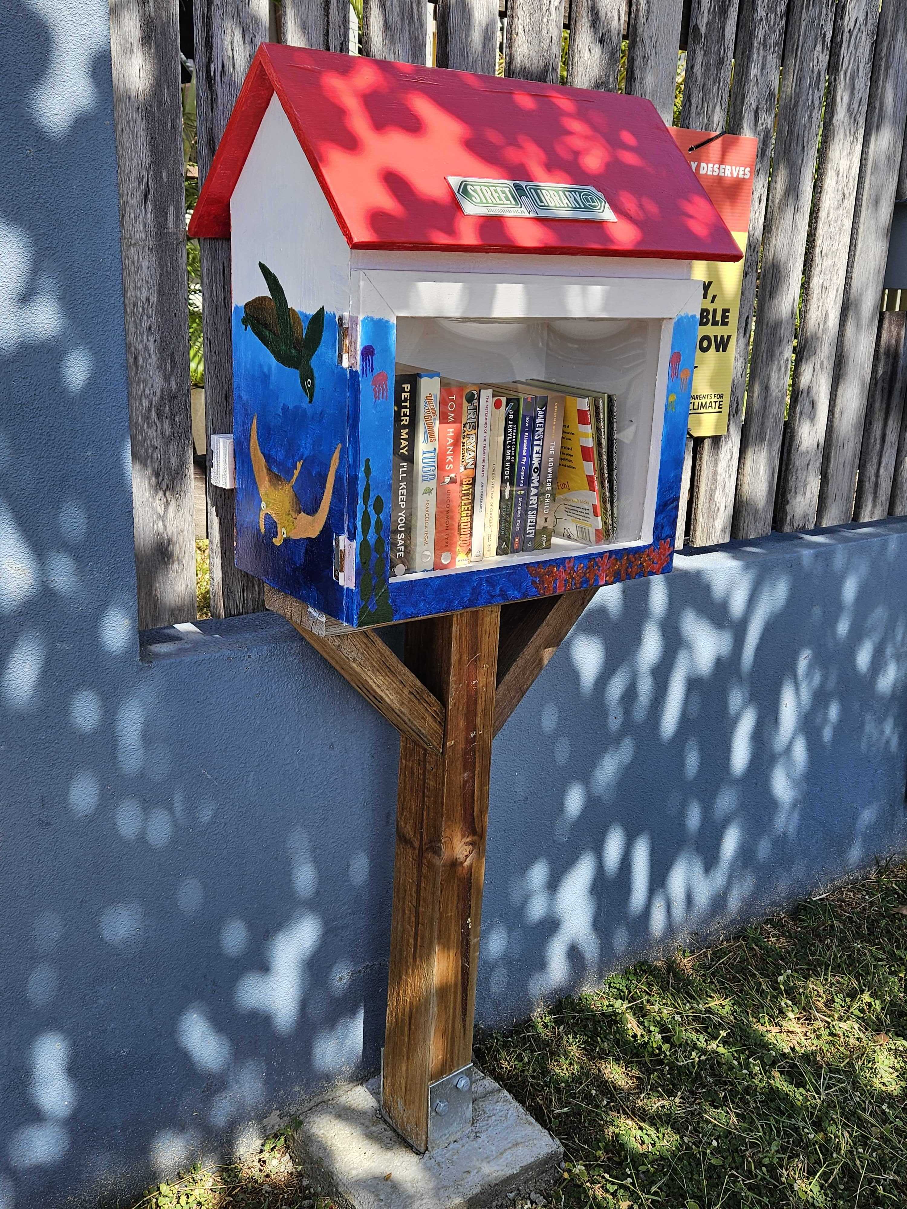 A colourful, marine-themed Little Street Library stands against NQCC's front fence