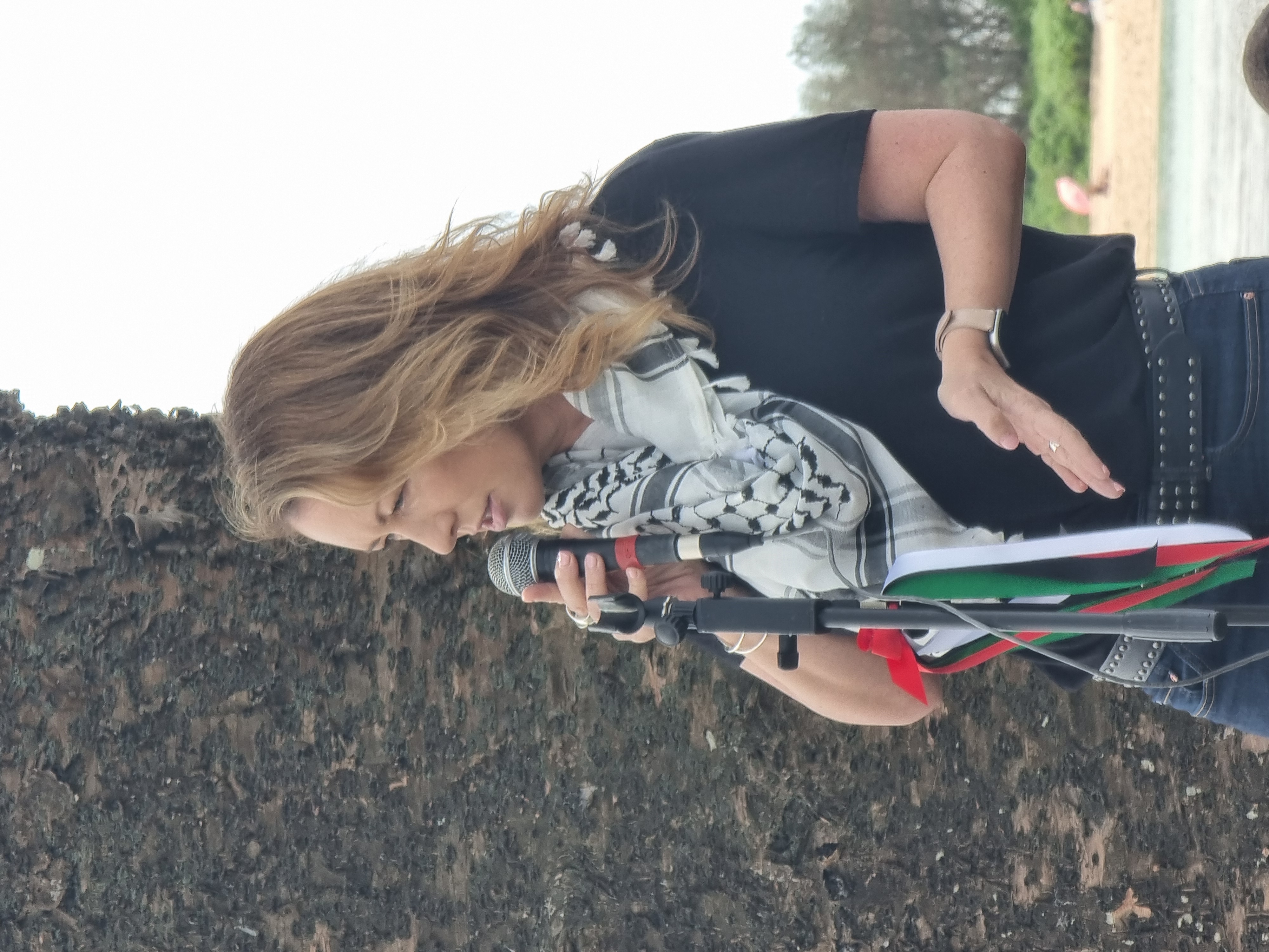 Photo of Abigail Boyd speaking at a rally, wearing a keffiyeh while standing and holding a microphone